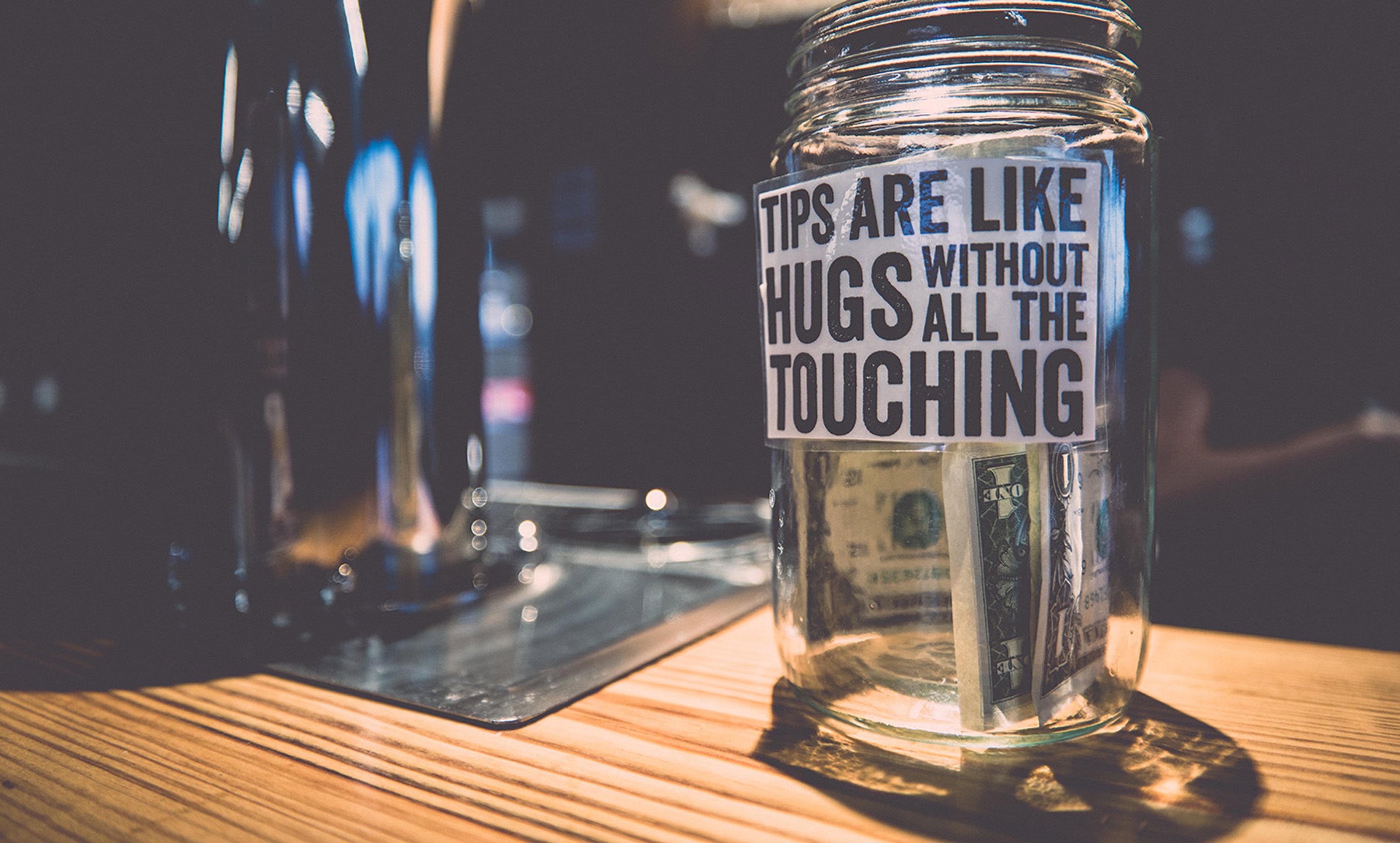 A tip jar on a wooden counter with a label that reads “Tips are like hugs without all the touching”, containing some banknotes.