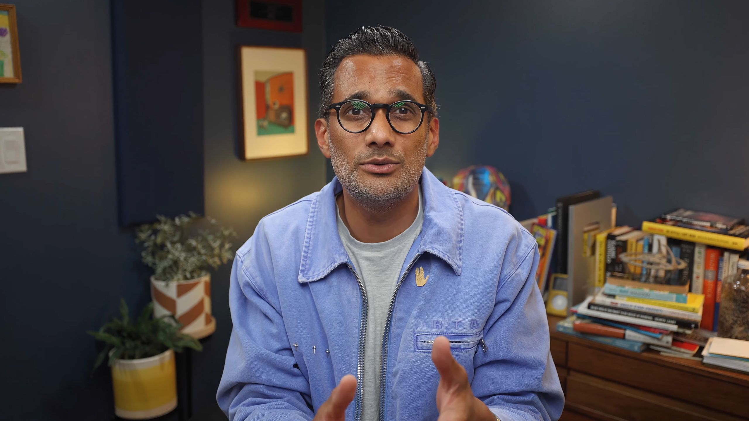 Photo of a man with glasses in a blue jacket speaking in a room with books, plants and colourful art in the background.