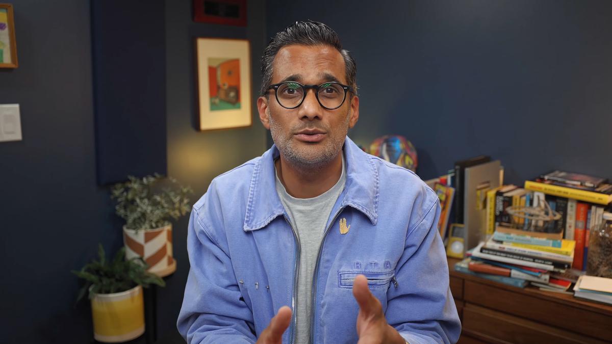 Photo of a man with glasses in a blue jacket speaking in a room with books, plants and colourful art in the background.