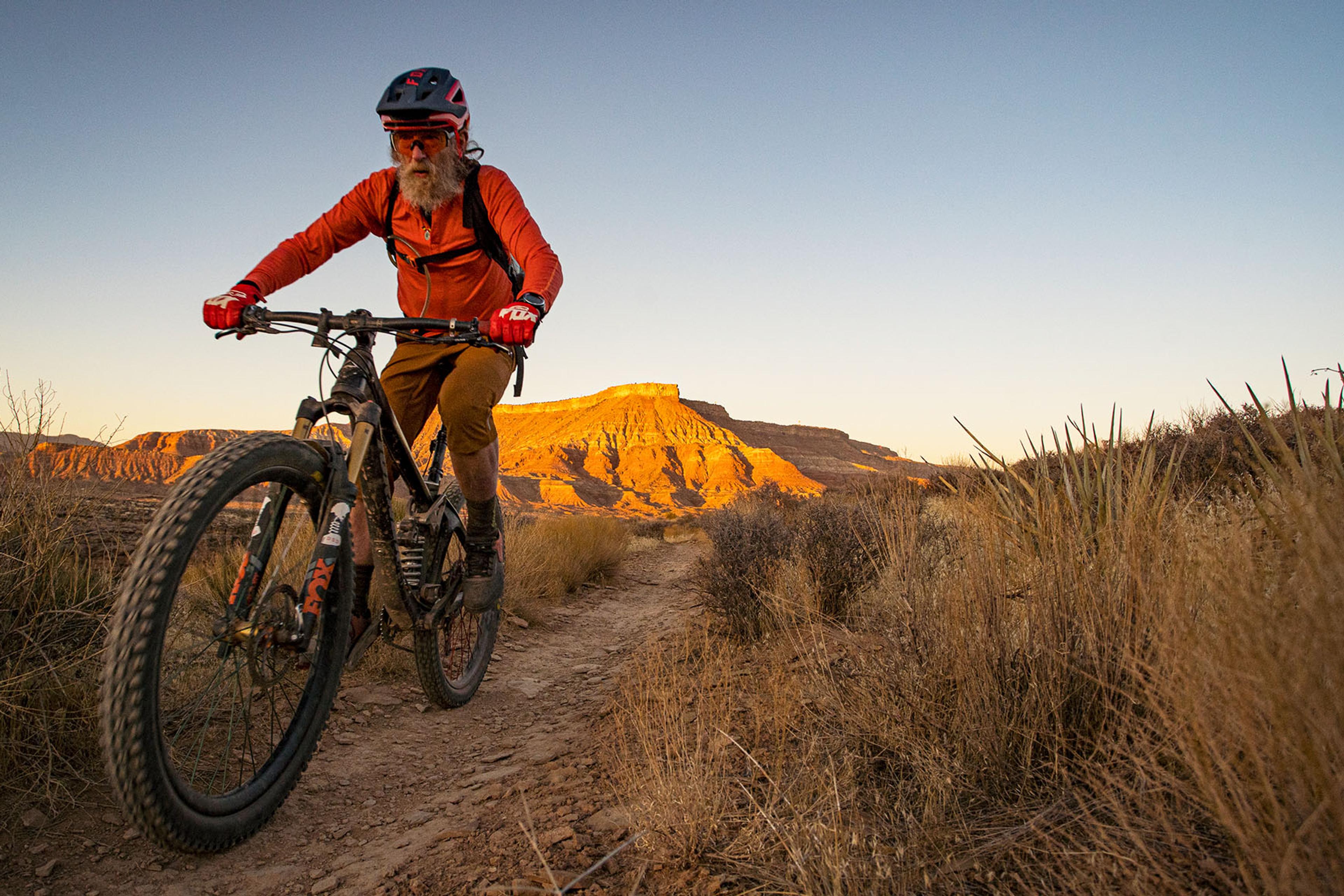 Mountain biker in orange gear riding at sunset on a dirt trail in a desert landscape with sandstone cliffs in the background.