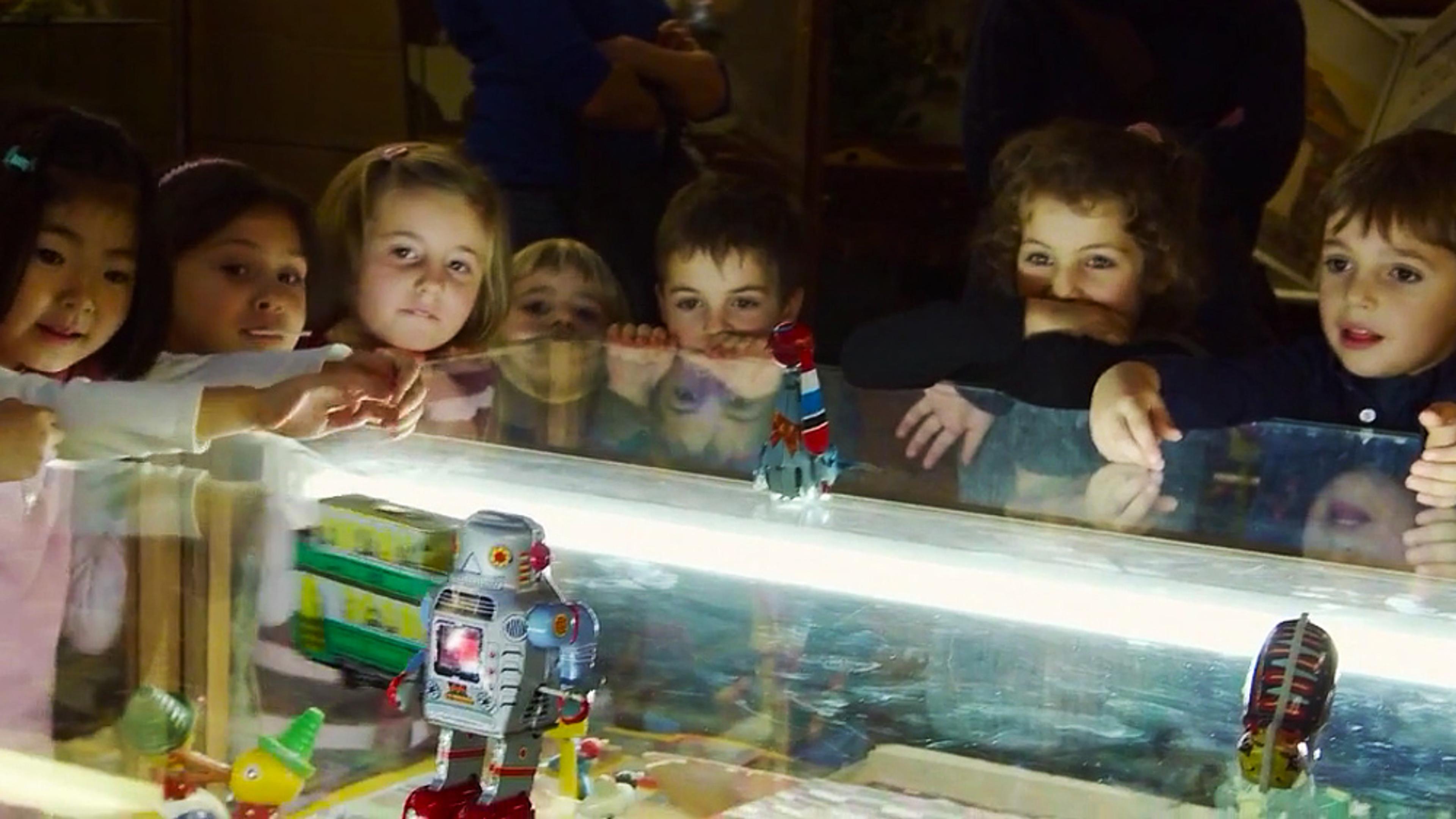 Children fascinated by a display of toy robots on a lit glass table in what appears to be a museum or exhibit room.