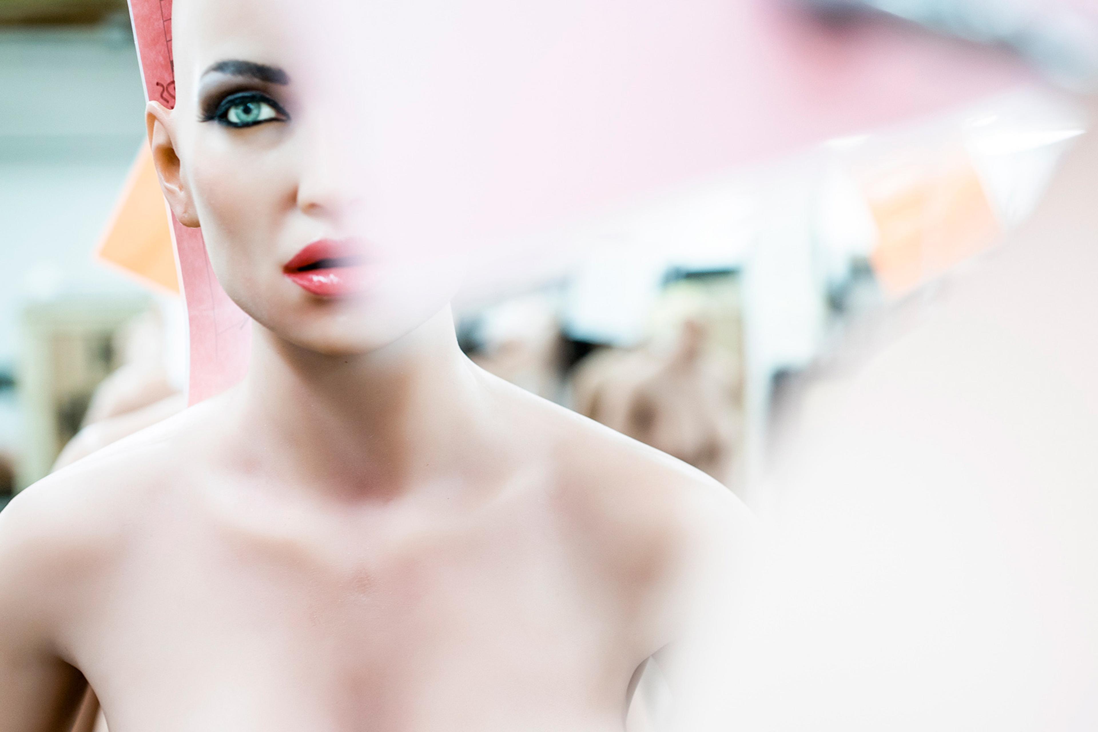 A mannequin head with painted blue eyes and red lips, partially obscured by a blurred pink and white shape in the foreground.