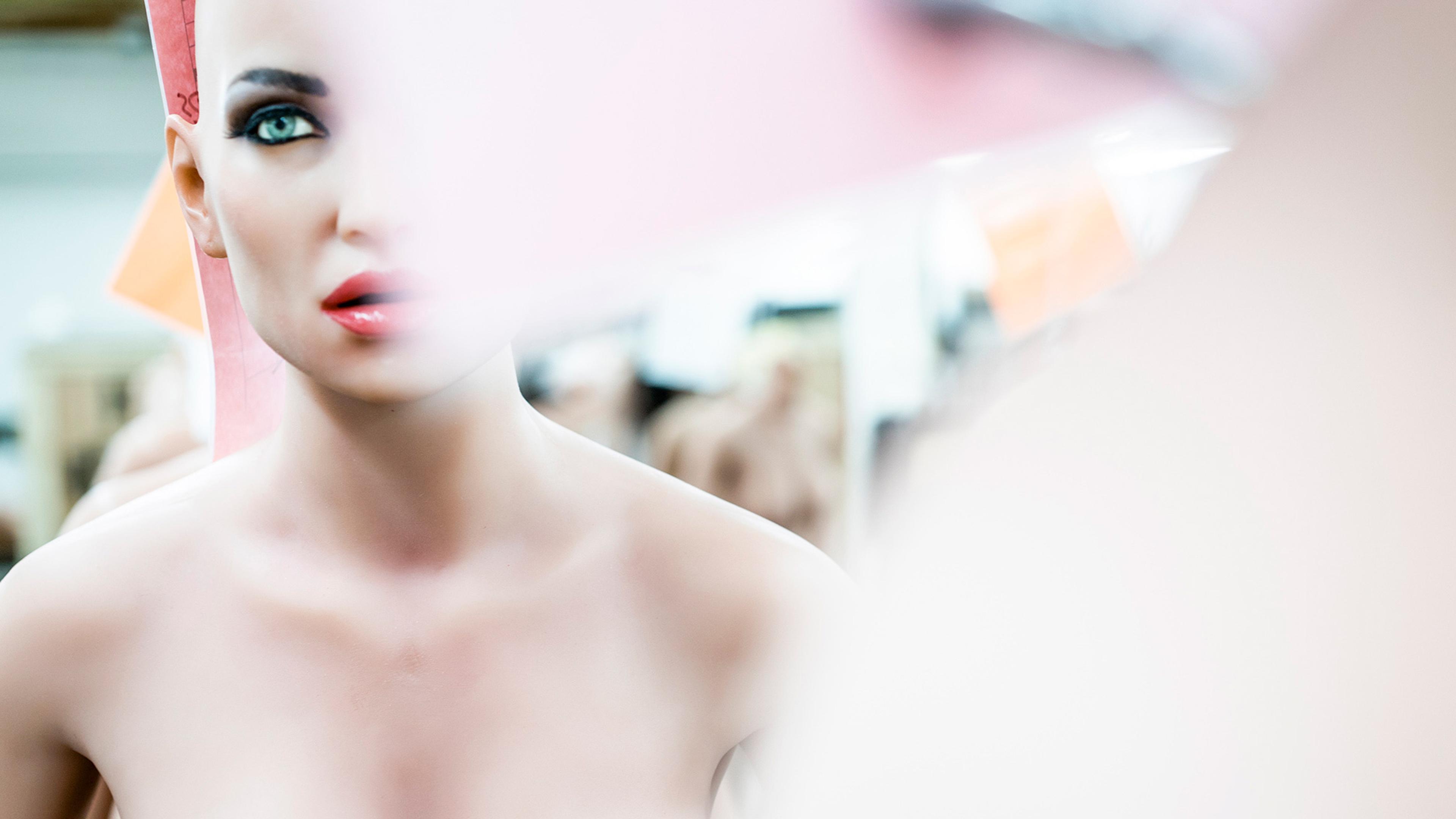 A mannequin head with painted blue eyes and red lips, partially obscured by a blurred pink and white shape in the foreground.