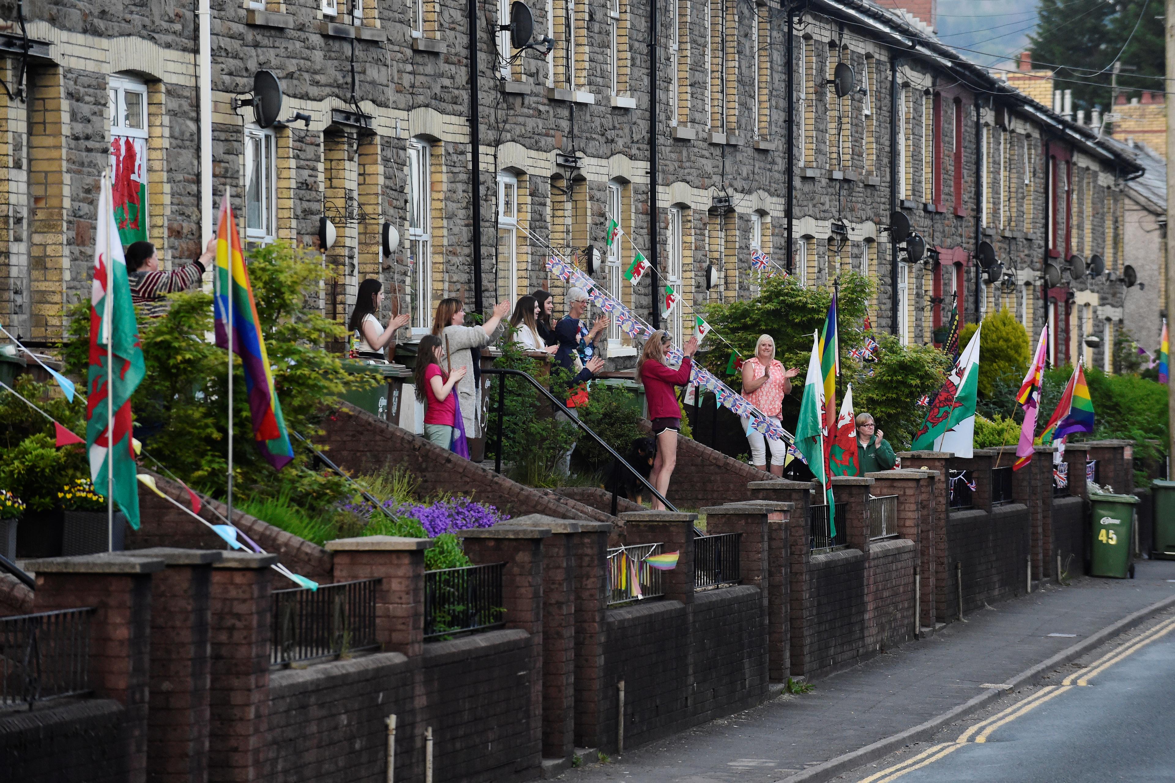 Photo of people clapping outside terraced houses decorated with flags and bunting, including Welsh flags and rainbow flags.