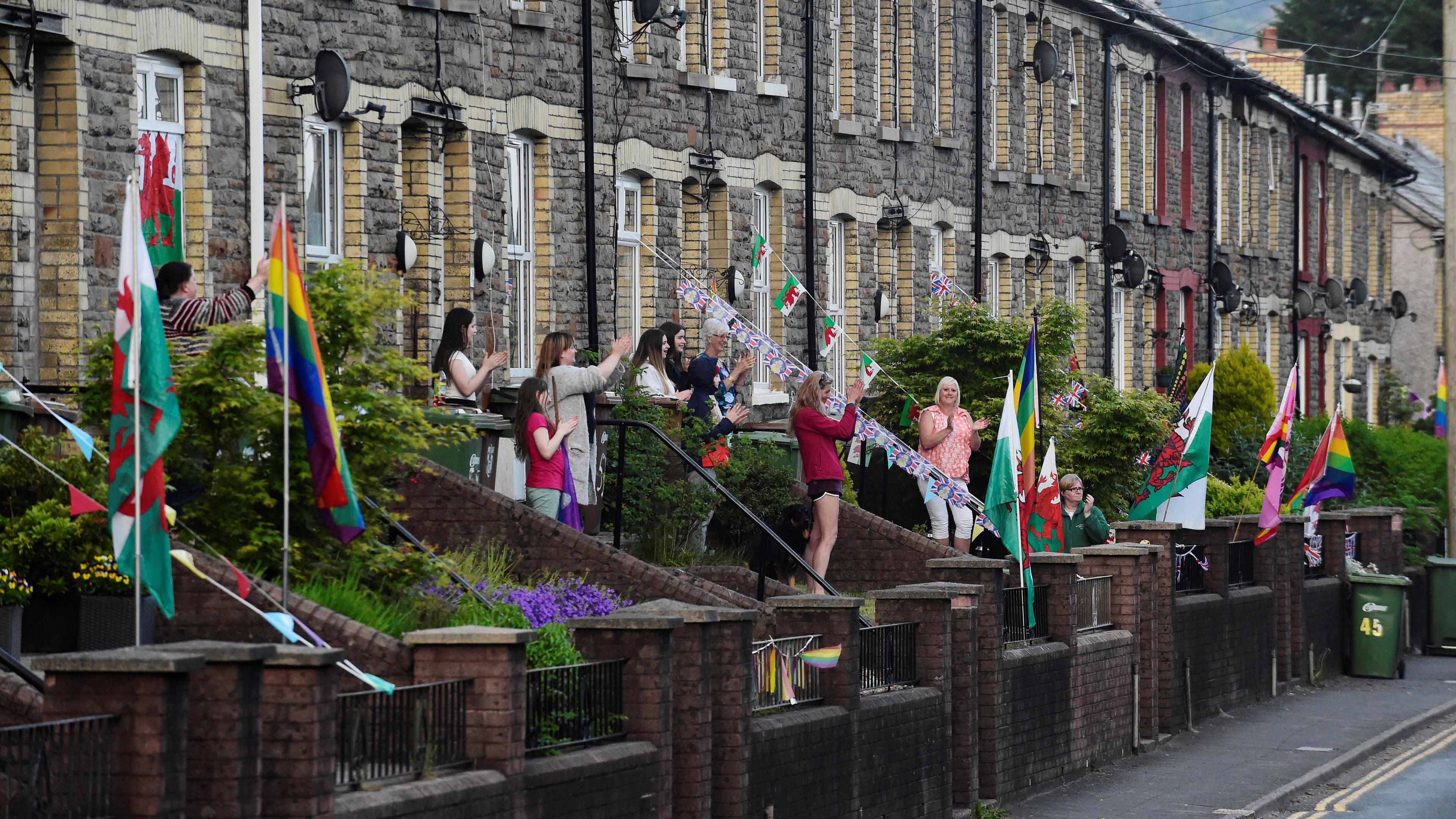 Photo of people clapping outside terraced houses decorated with flags and bunting, including Welsh flags and rainbow flags.