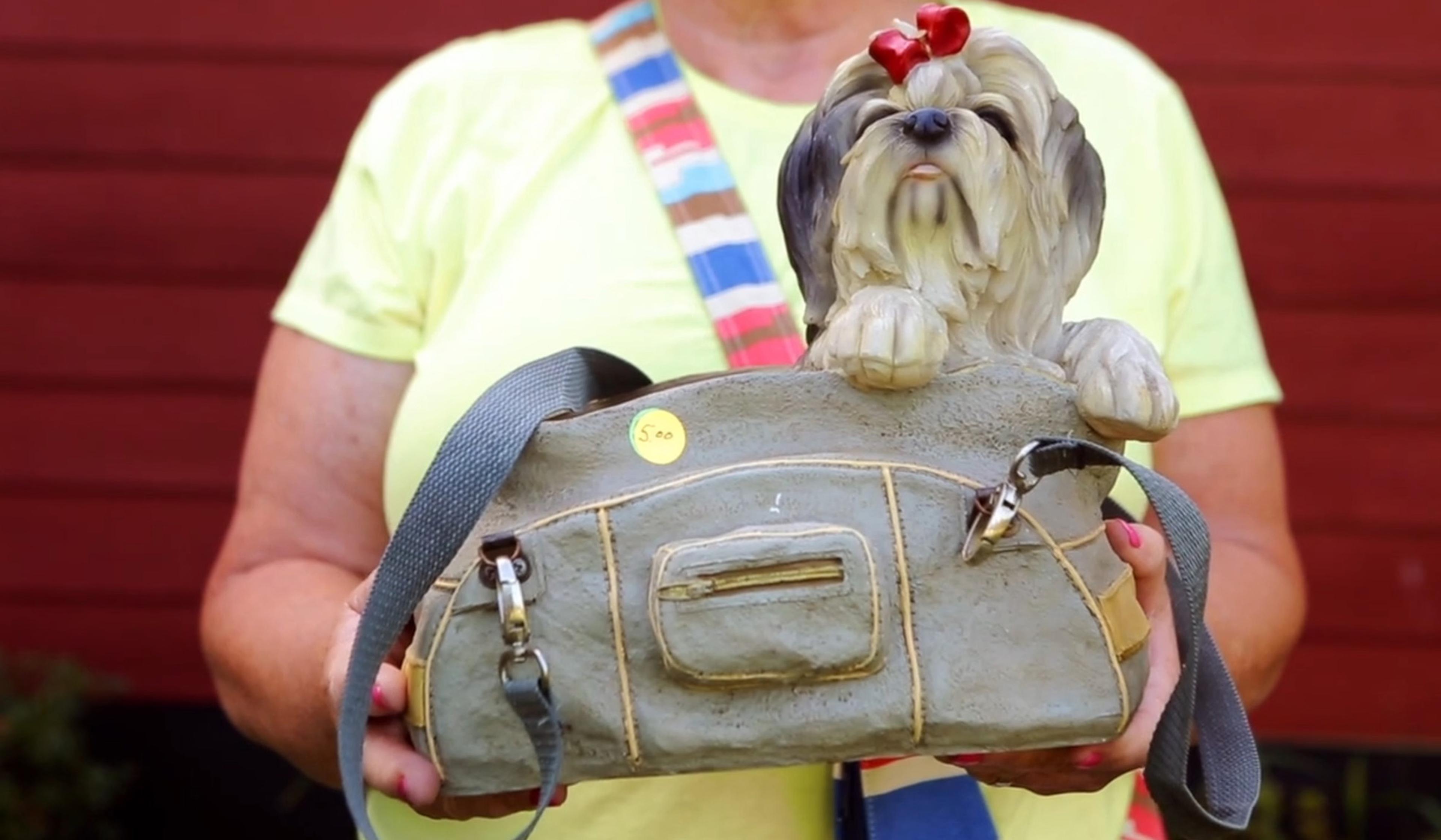 A person holding a figurine of a grey Shih Tzu dog wearing a red bow in a handbag with a $5 price sticker on
