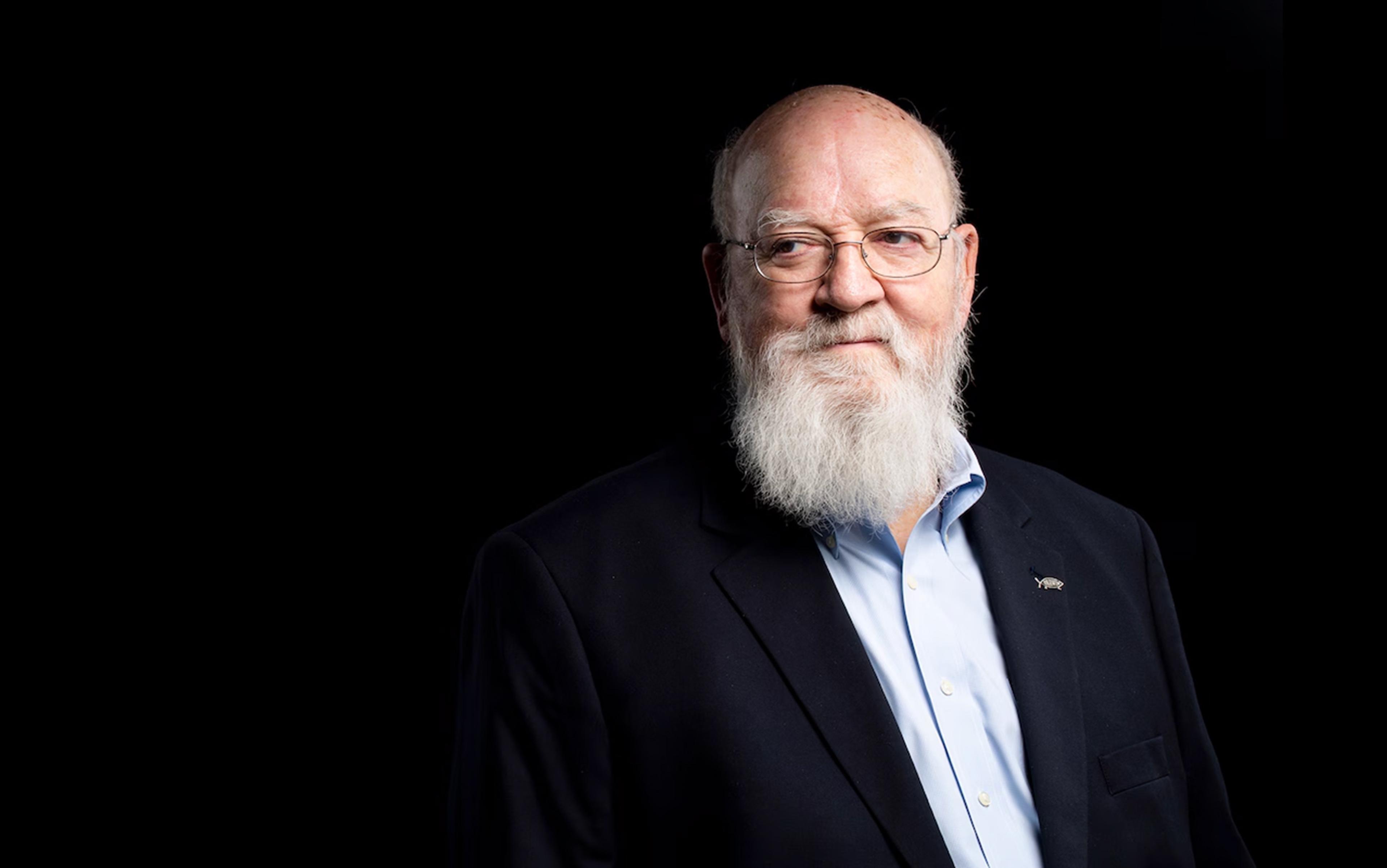 Photo of Daniel Dennett looking to the side with a white beard and glasses wearing a suit against a black background.