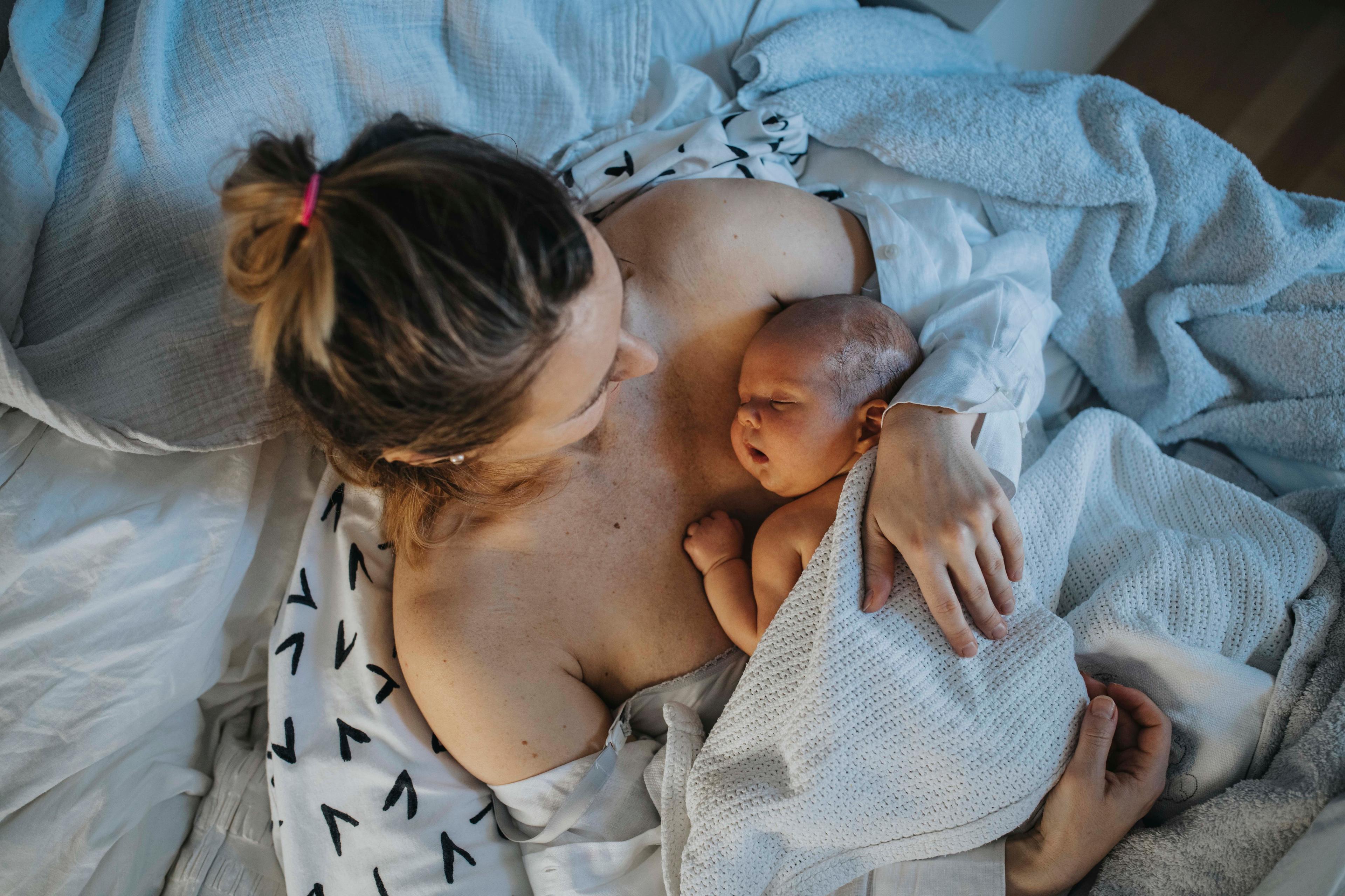 Photo of a mother lying in bed holding her newborn baby, both wrapped in white blankets, showing tenderness and care.
