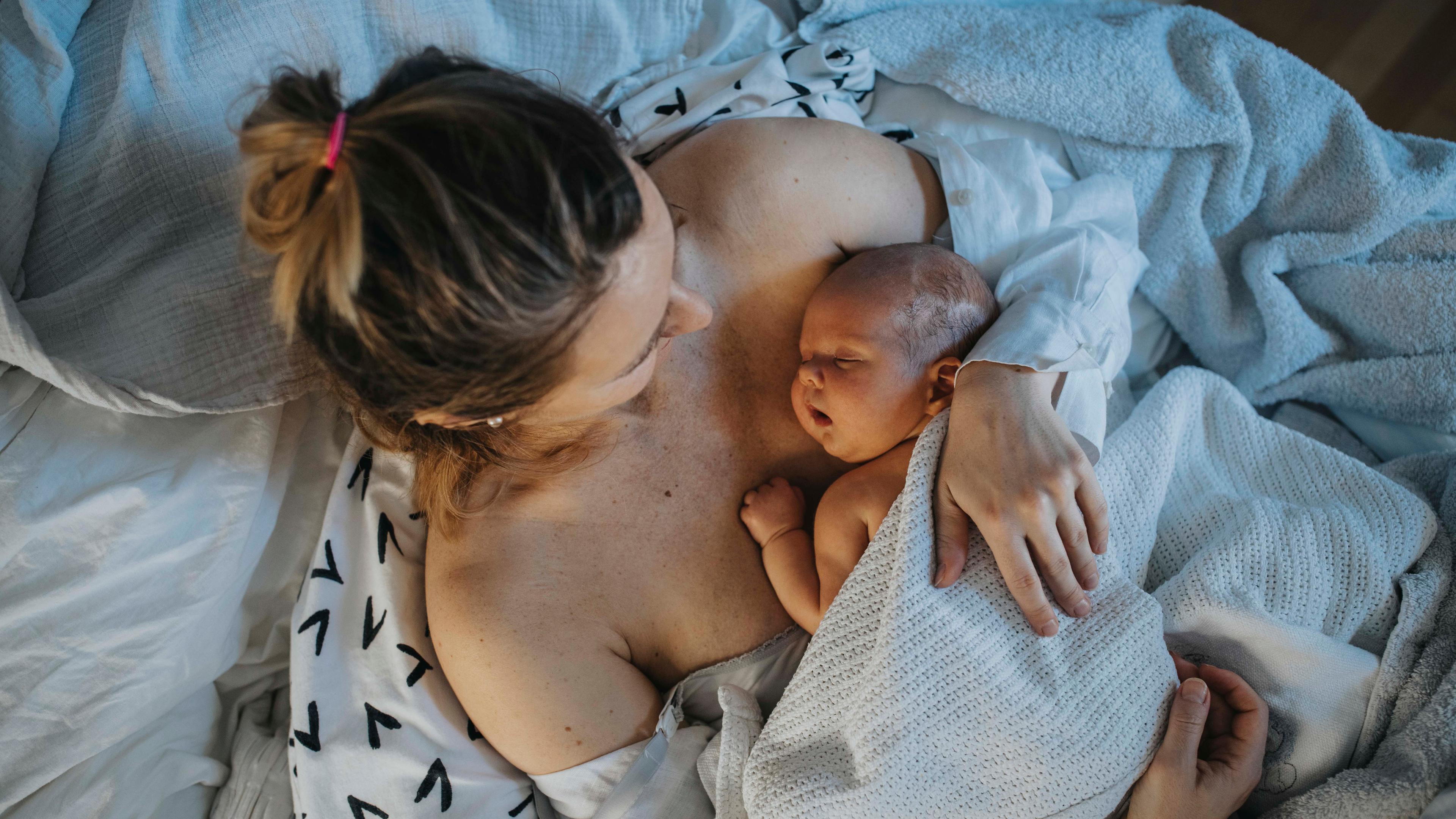 Photo of a mother lying in bed holding her newborn baby, both wrapped in white blankets, showing tenderness and care.