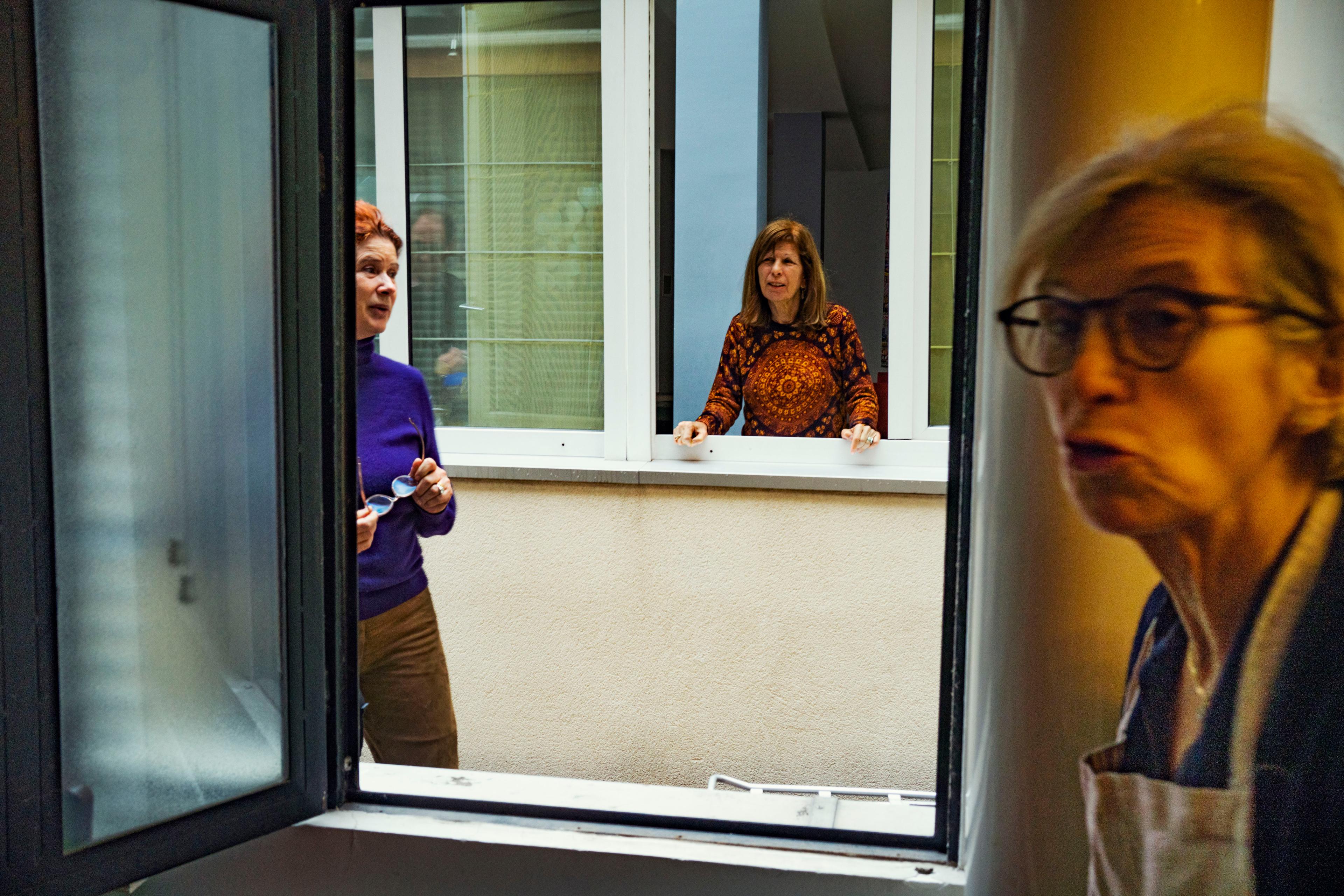 Photo of three women talking through open windows, showing interaction between apartments in a residential building.