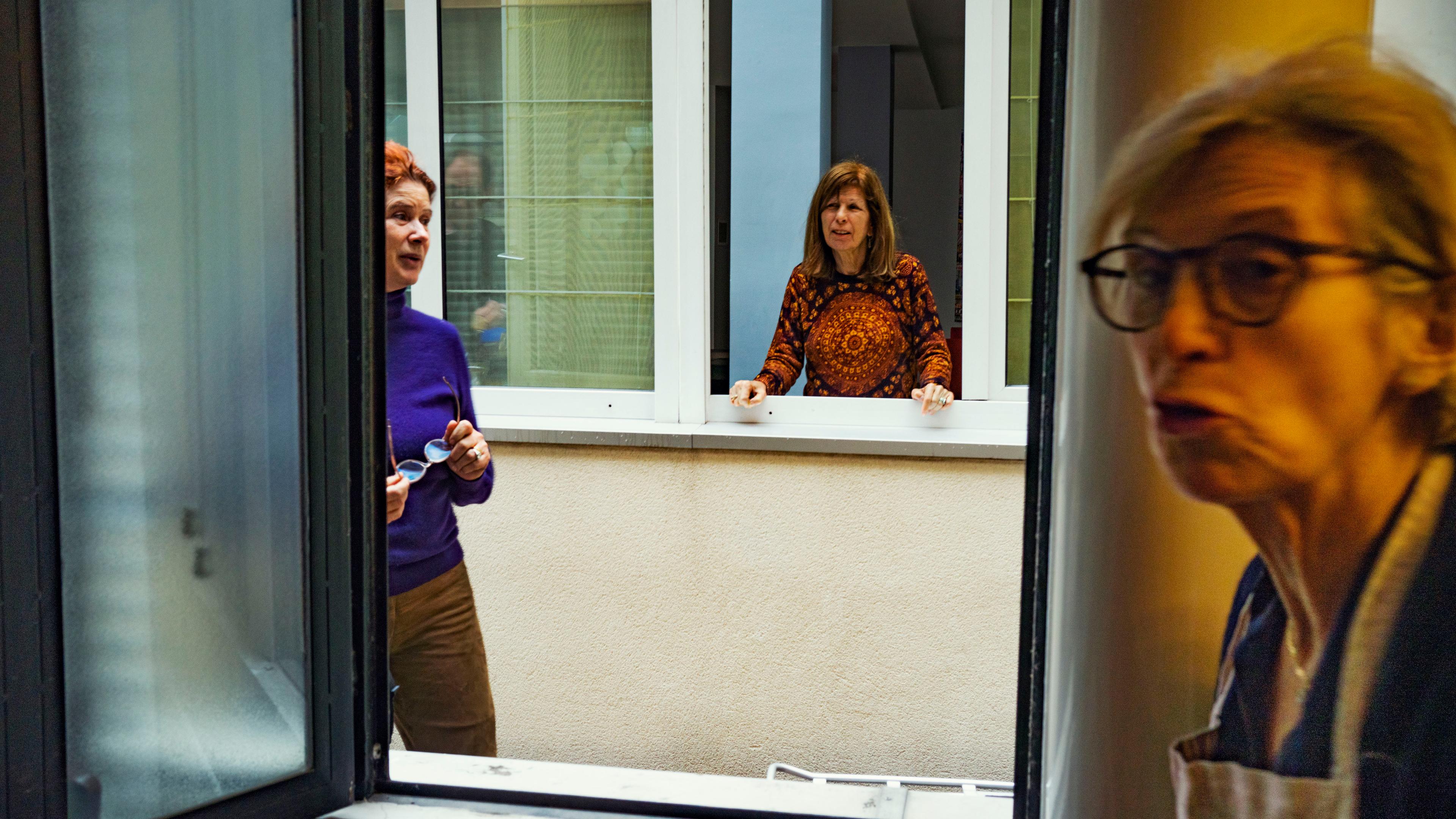 Photo of three women talking through open windows, showing interaction between apartments in a residential building.
