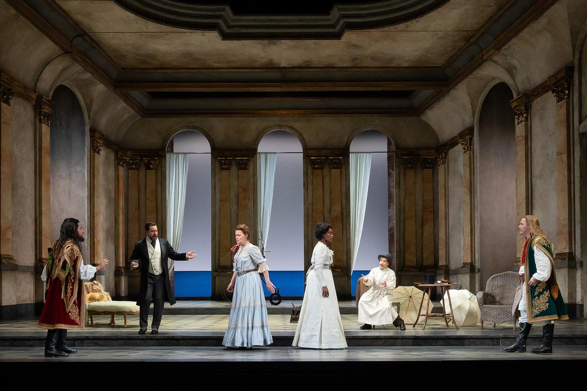 A theatrical stage with six actors in historical costumes set in a grand hall with arches and curtains.