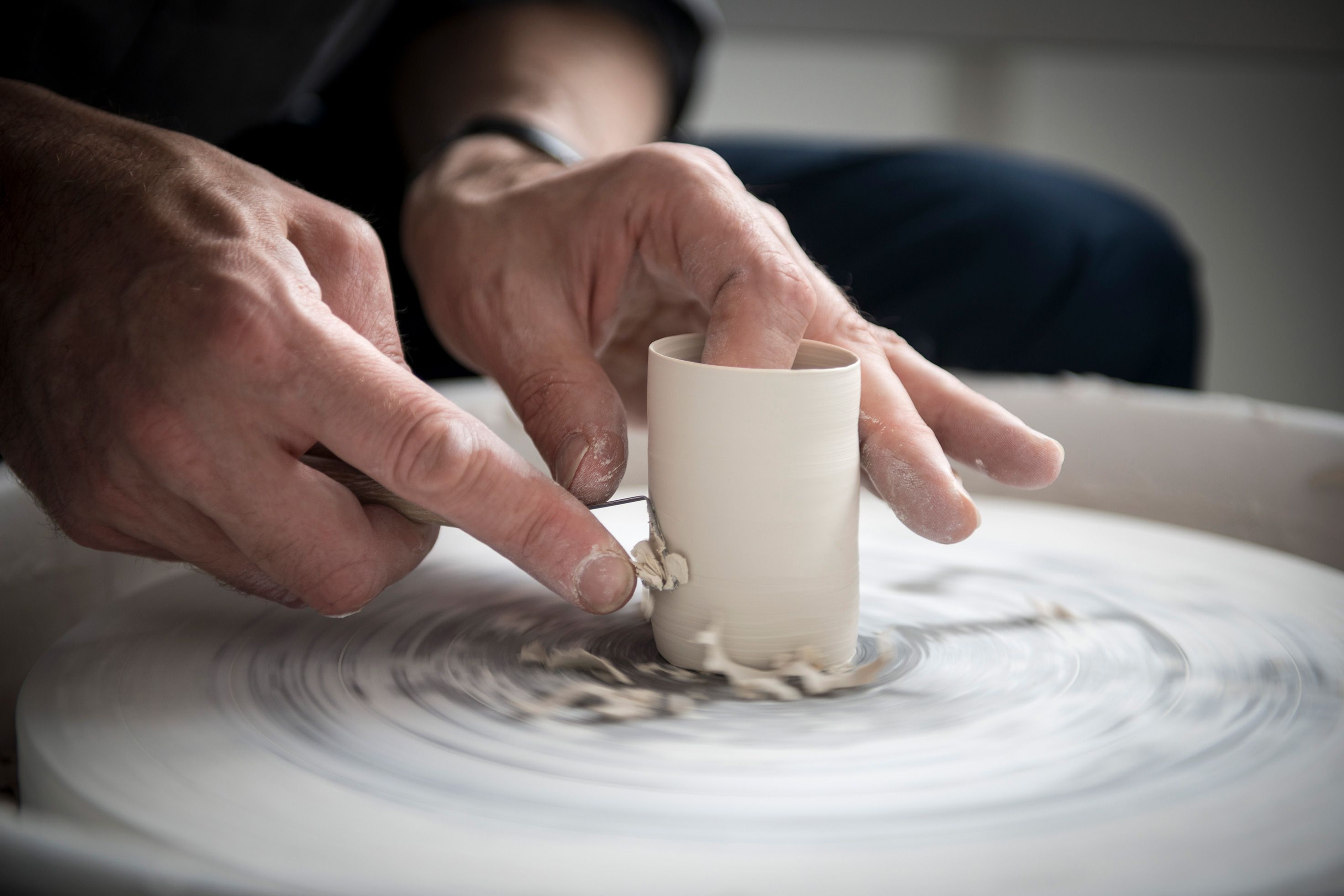 Hands shaping clay on a spinning pottery wheel, with a tool used for carving.