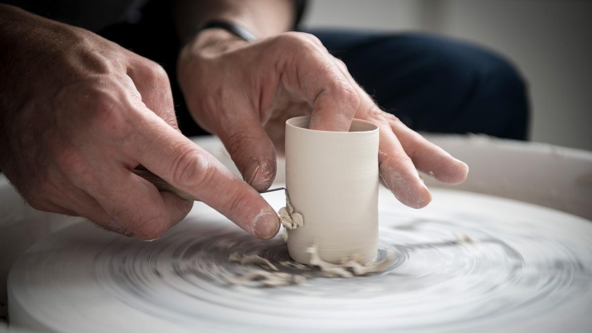 Hands shaping clay on a spinning pottery wheel, with a tool used for carving.
