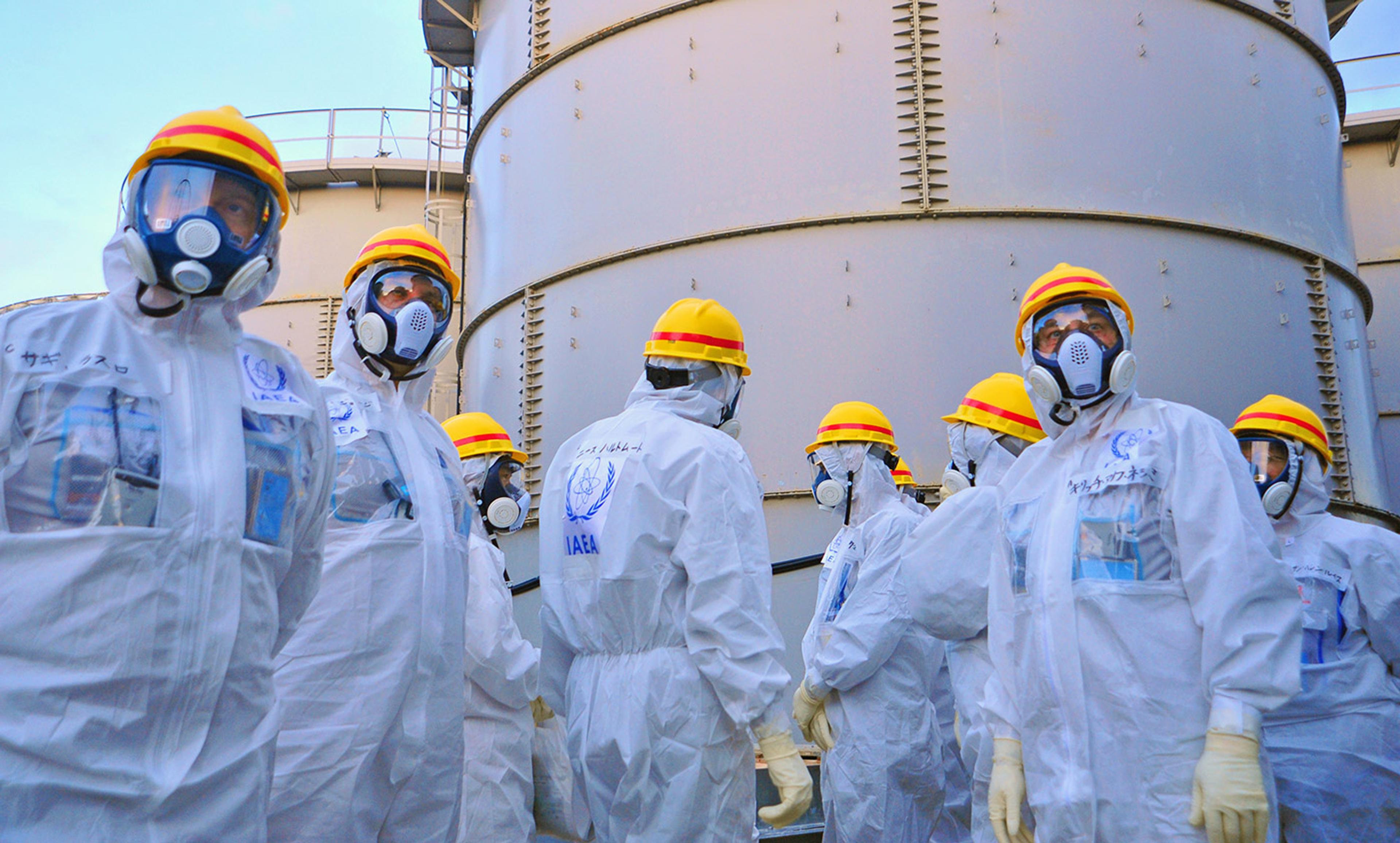 Workers in white hazmat suits, yellow helmets and face masks at an industrial site with large cylindrical structures in the background.