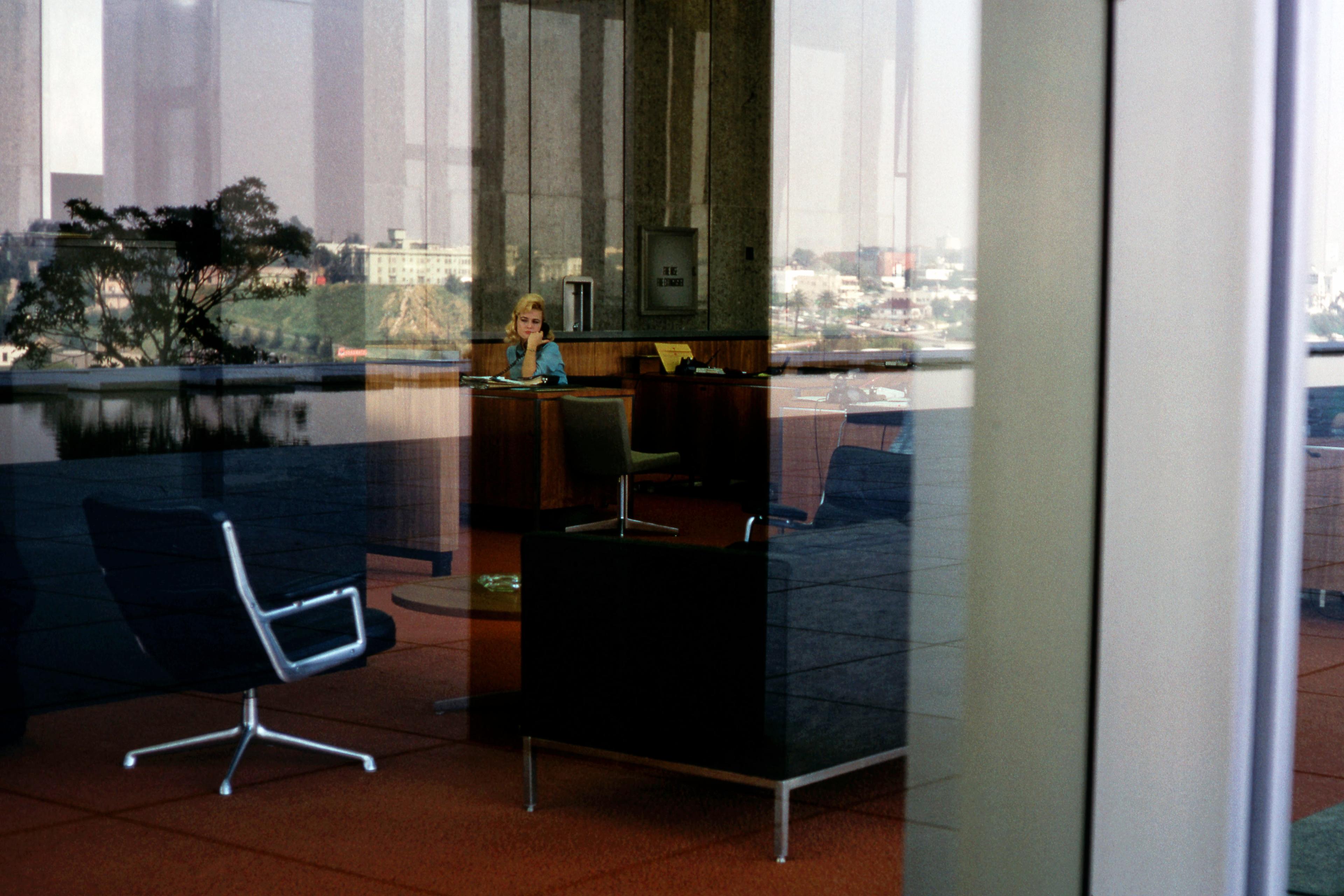 Photo of an office viewed through glass showing a woman at a desk, cityscape reflected outside.