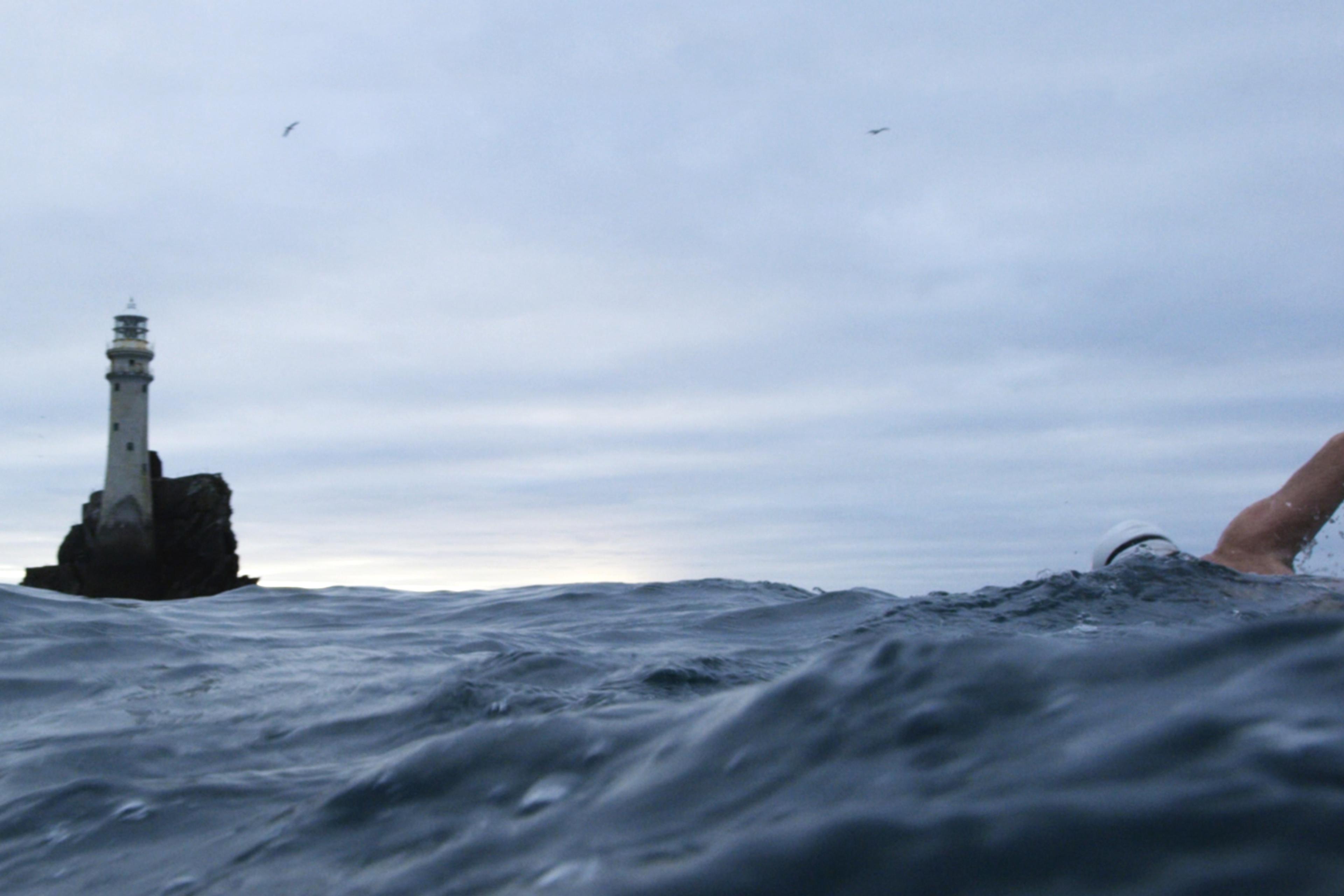 A lone swimmer in choppy waters near a lighthouse on a rocky outcrop, under an overcast sky.