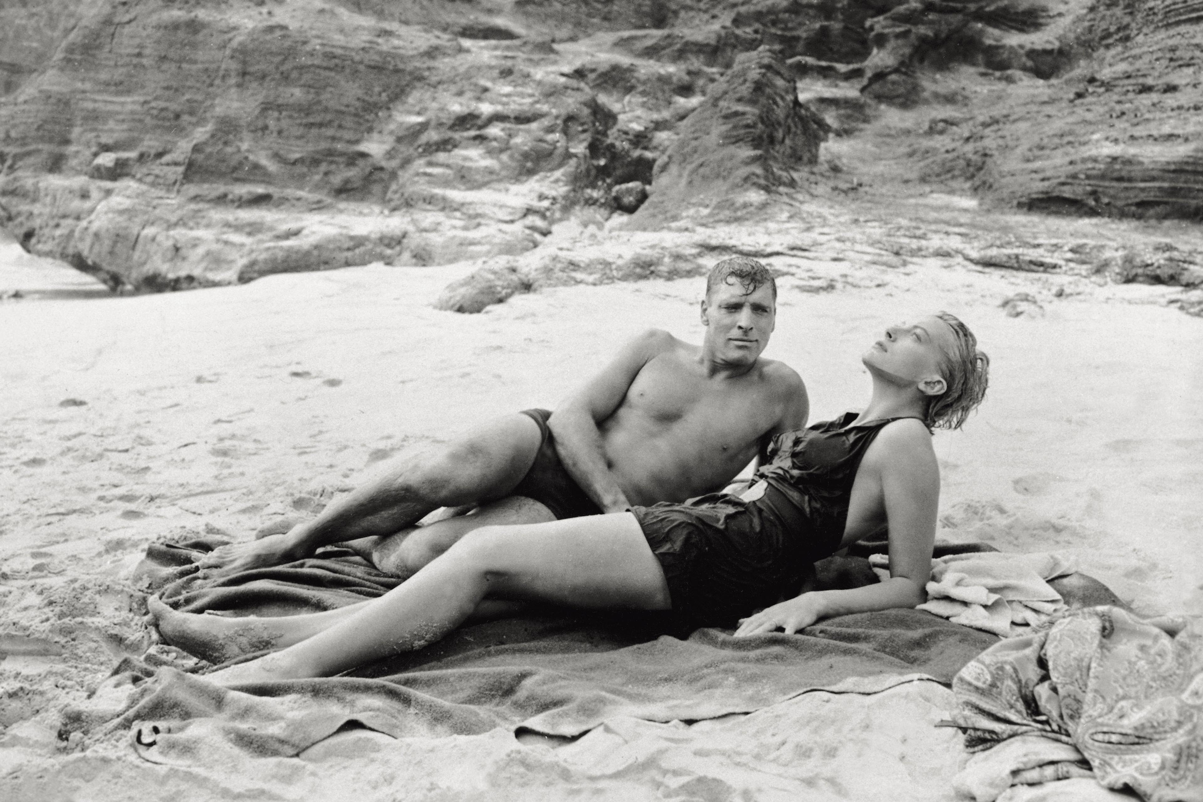 Black and white photo of a man and woman in swimwear relaxing on a sandy beach with rocky cliffs in the background.