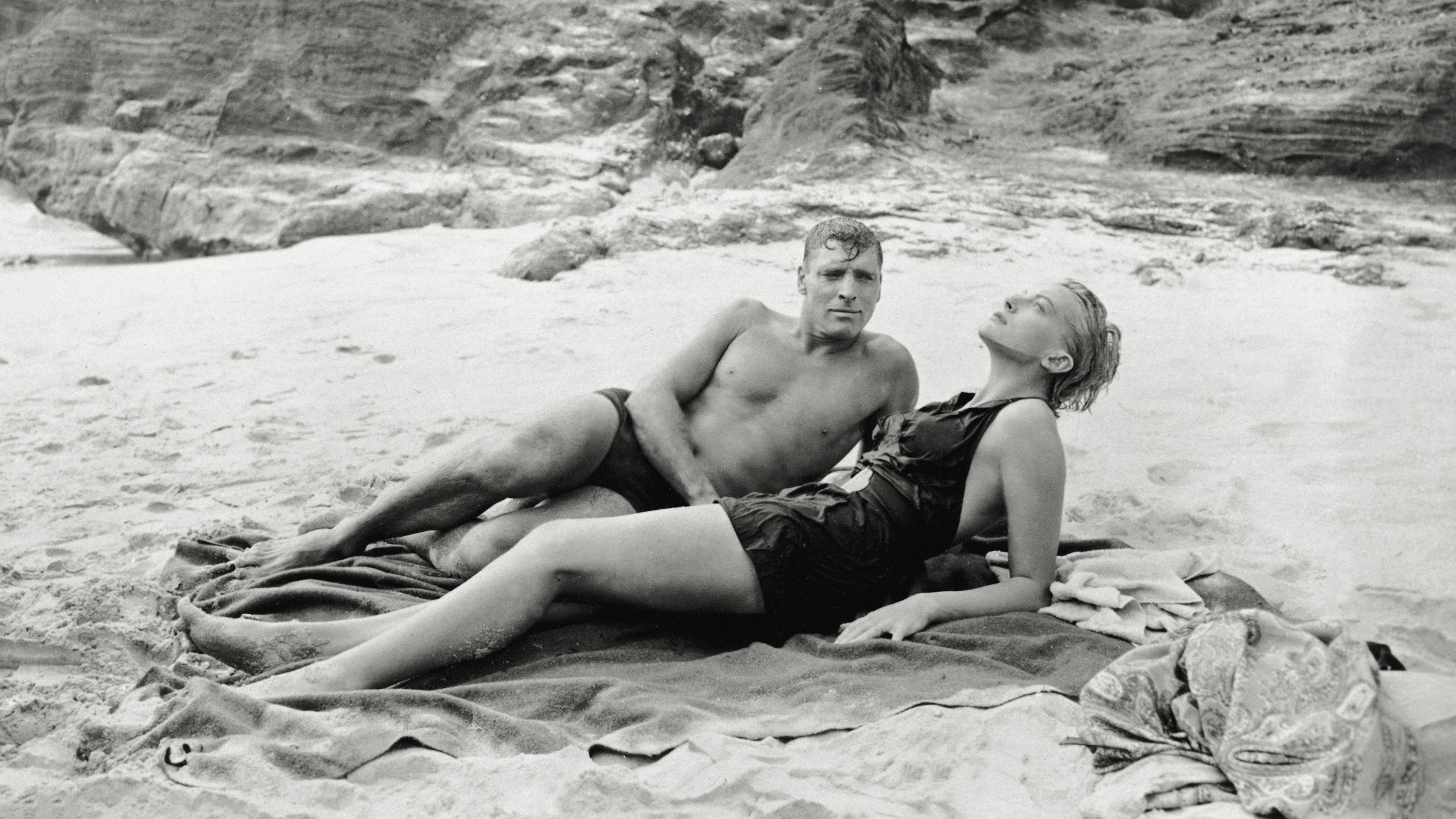 Black and white photo of a man and woman in swimwear relaxing on a sandy beach with rocky cliffs in the background.