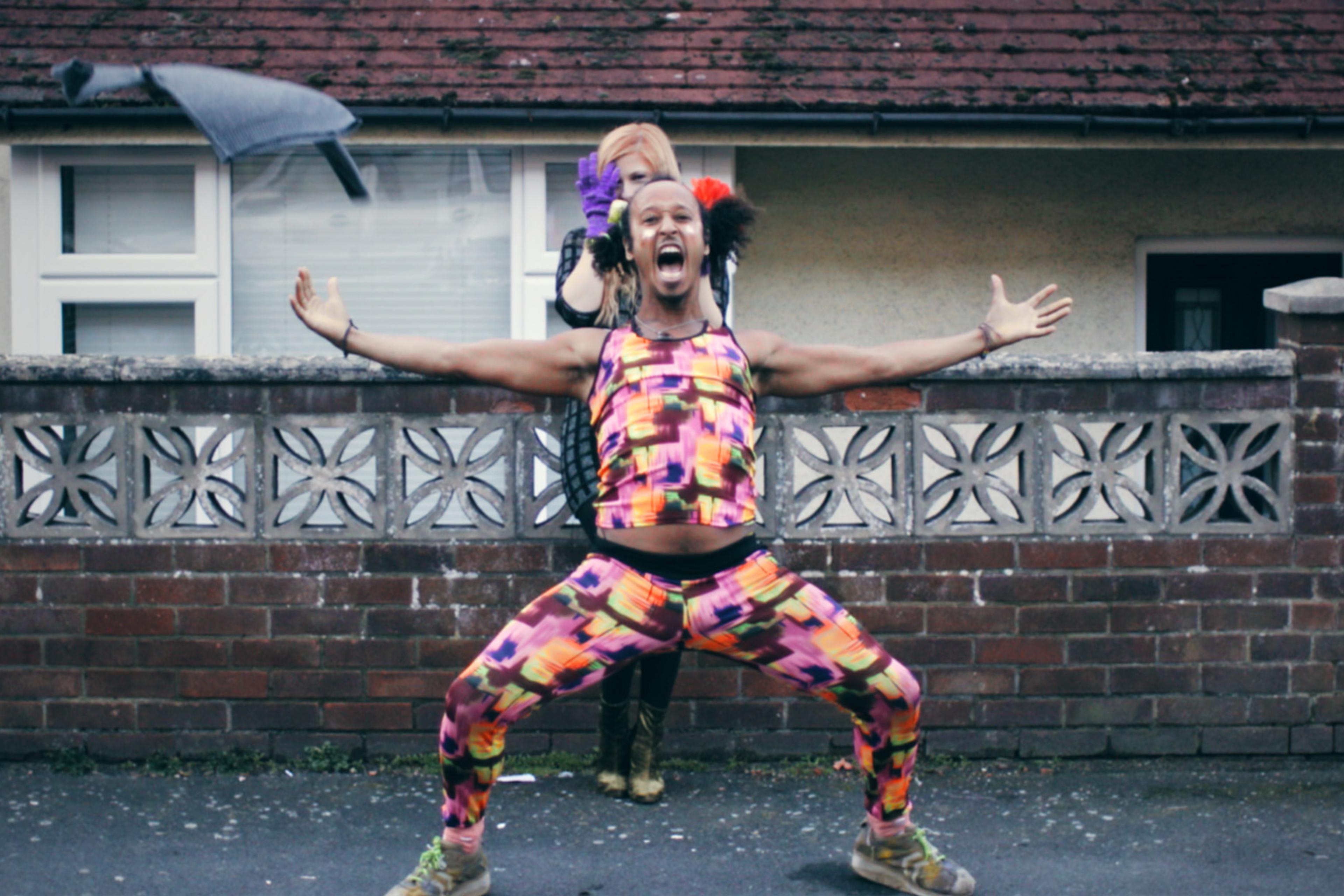 A person in vibrant, multicoloured activewear energetically poses with arms outstretched, while another stands behind. A jumper is mid-air above them.