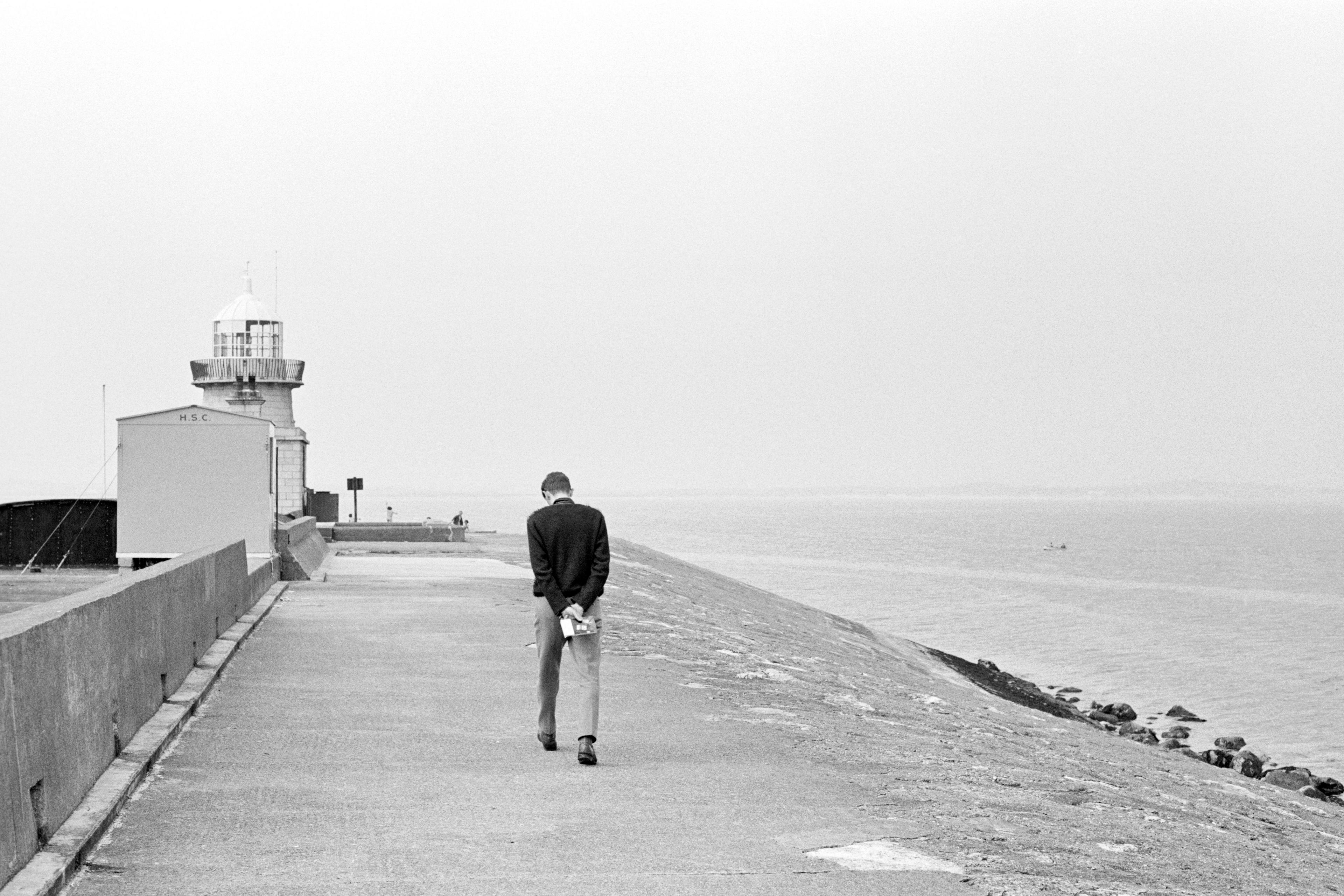 Black and white photo of a man walking towards a lighthouse by the sea with his hands behind his back.