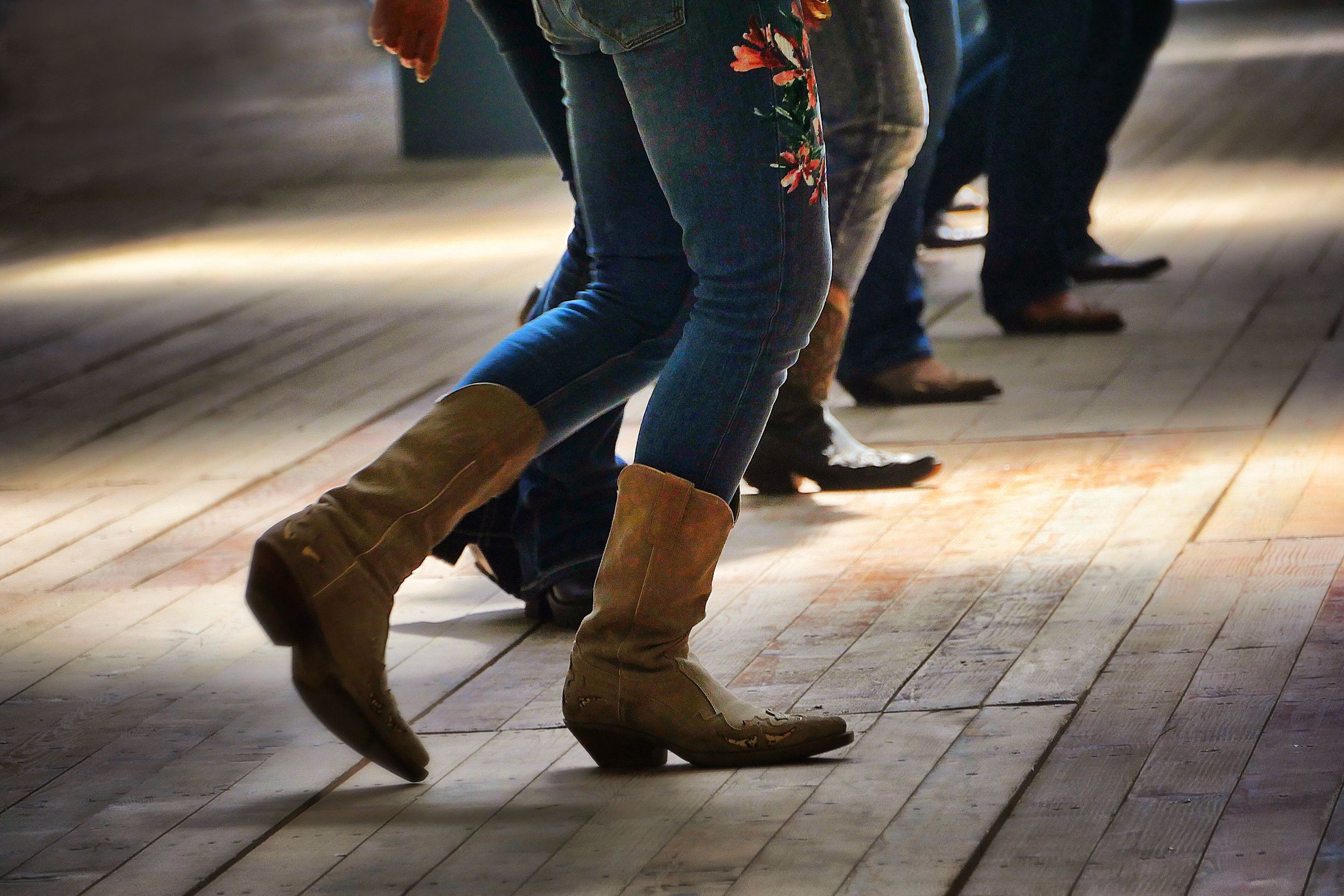 People line dancing indoors, wearing jeans and boots on a wooden floor.