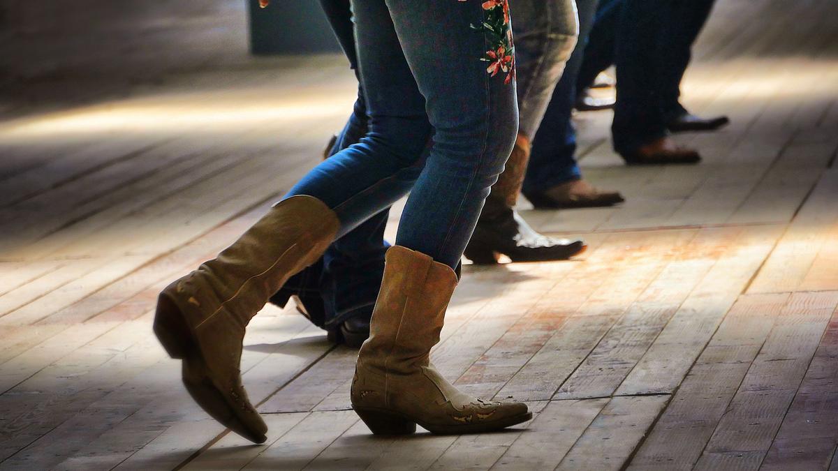 People line dancing indoors, wearing jeans and boots on a wooden floor.