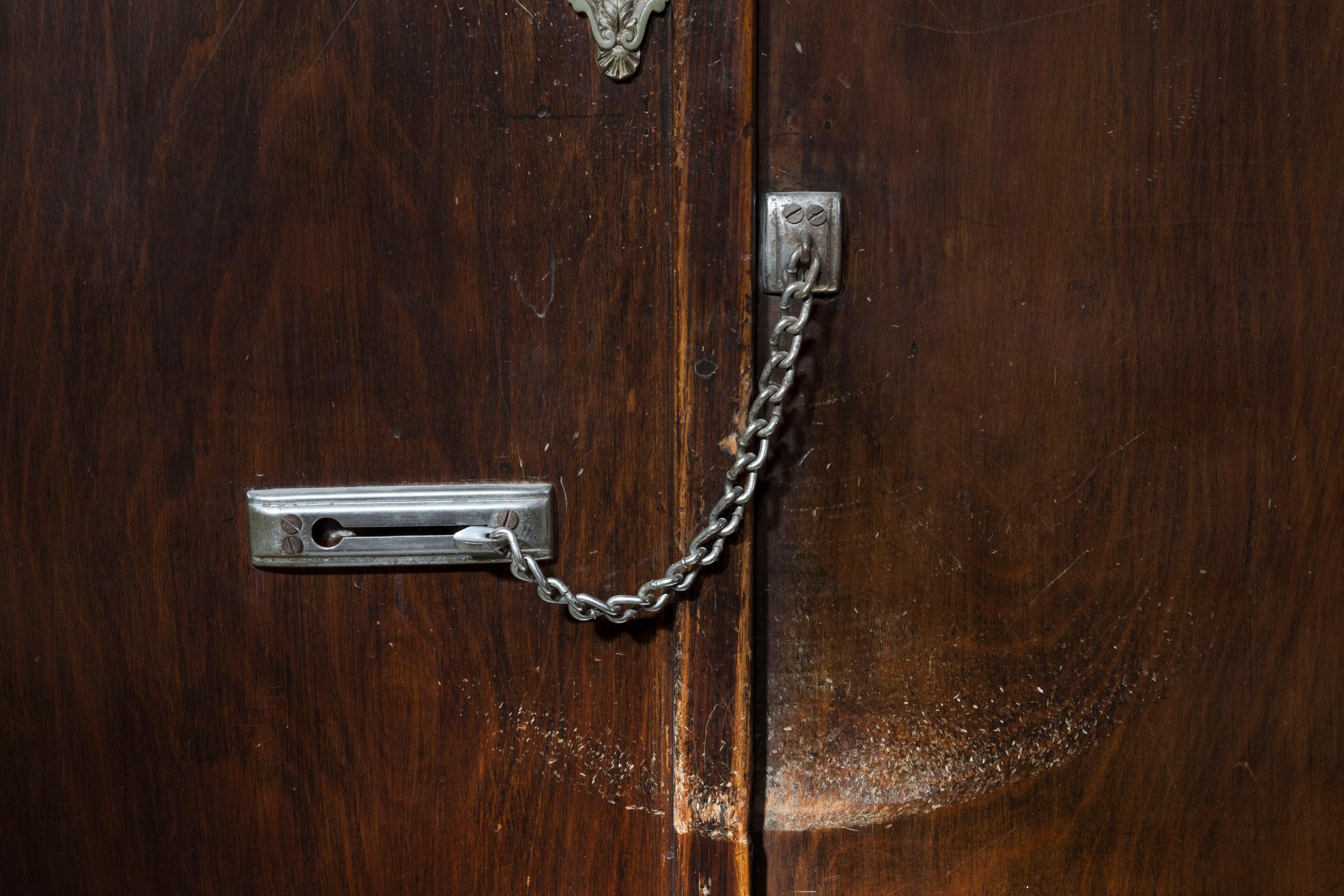 Photo of a wooden door with a metal chain lock, slightly scuffed surface and ornate latch on top against a dark background.