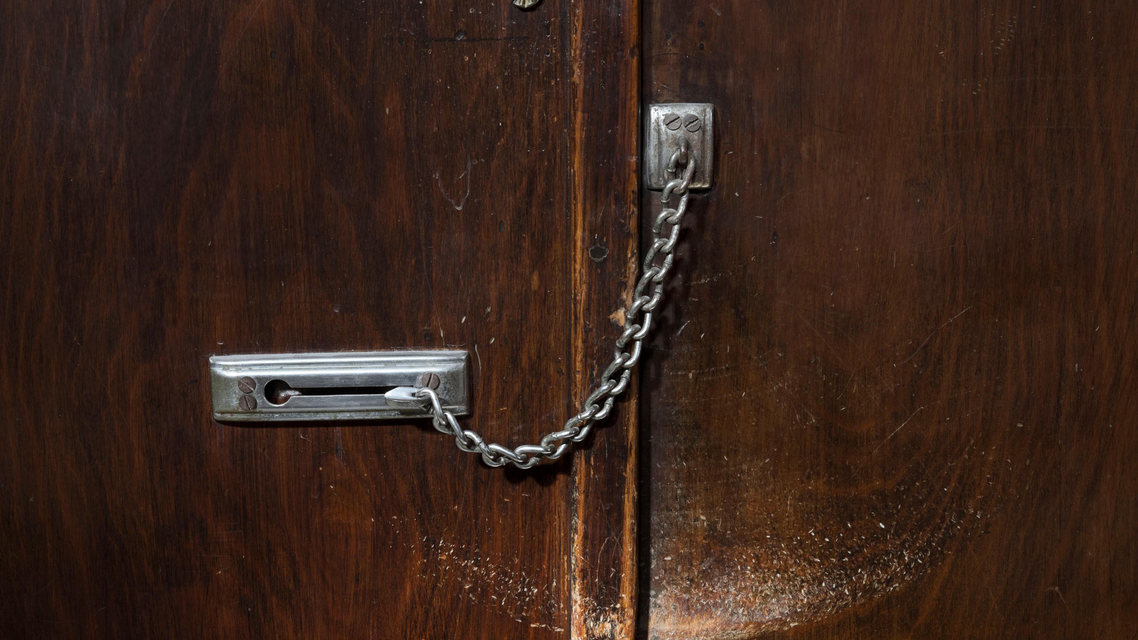 Photo of a wooden door with a metal chain lock, slightly scuffed surface and ornate latch on top against a dark background.