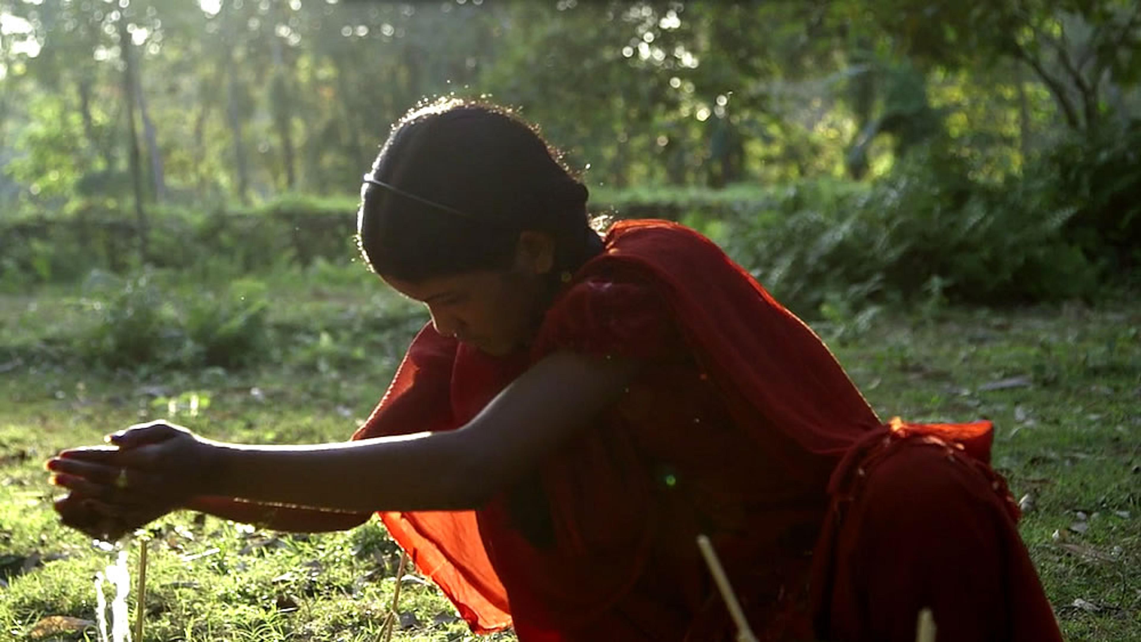 A person in a red garment bent over with hands extended forward, outdoors in a lush green area with sunlight filtering through.