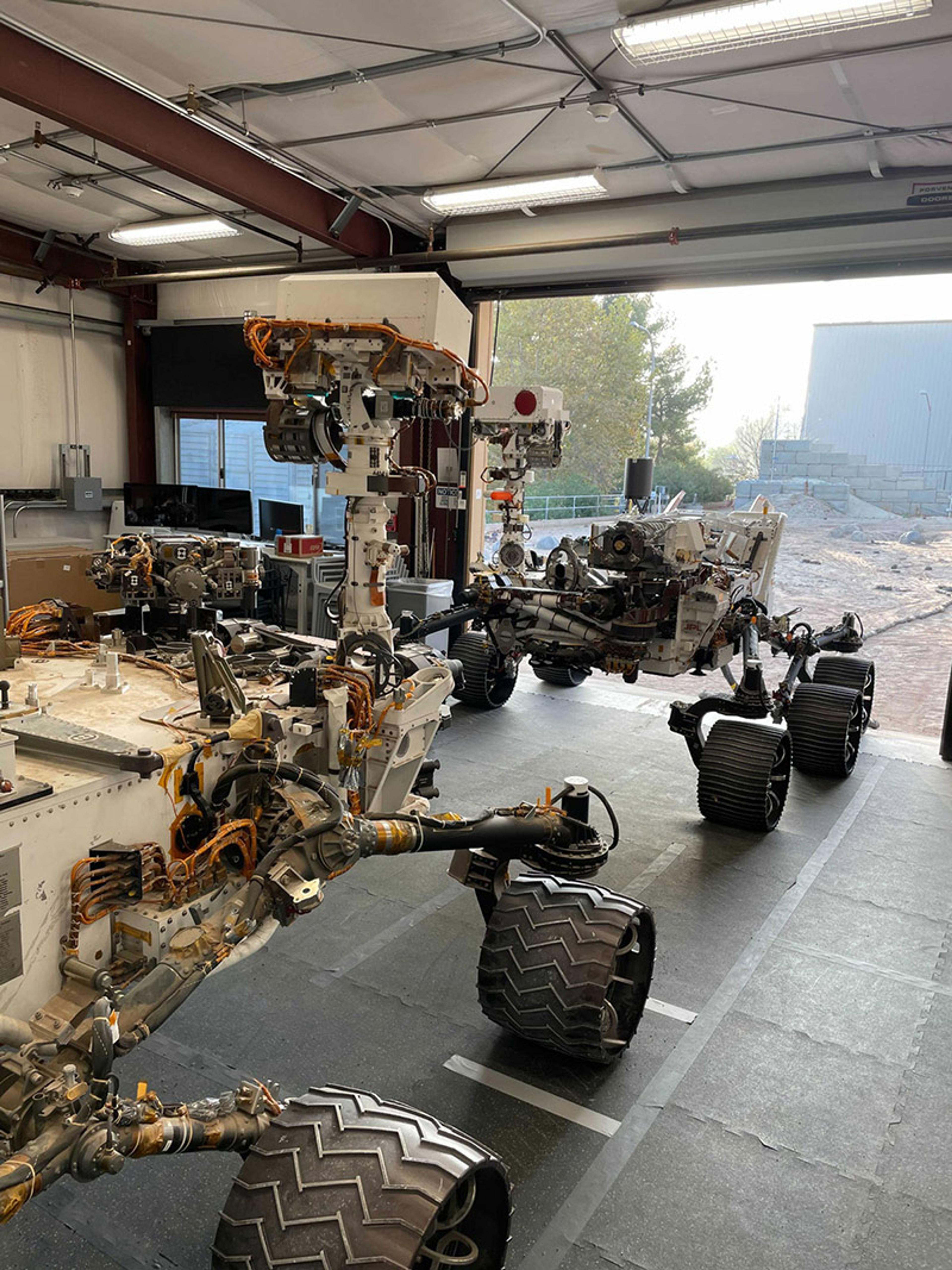 Two Mars rovers in a workshop with various equipment, cables, and large wheels, seen from an angle inside the building.