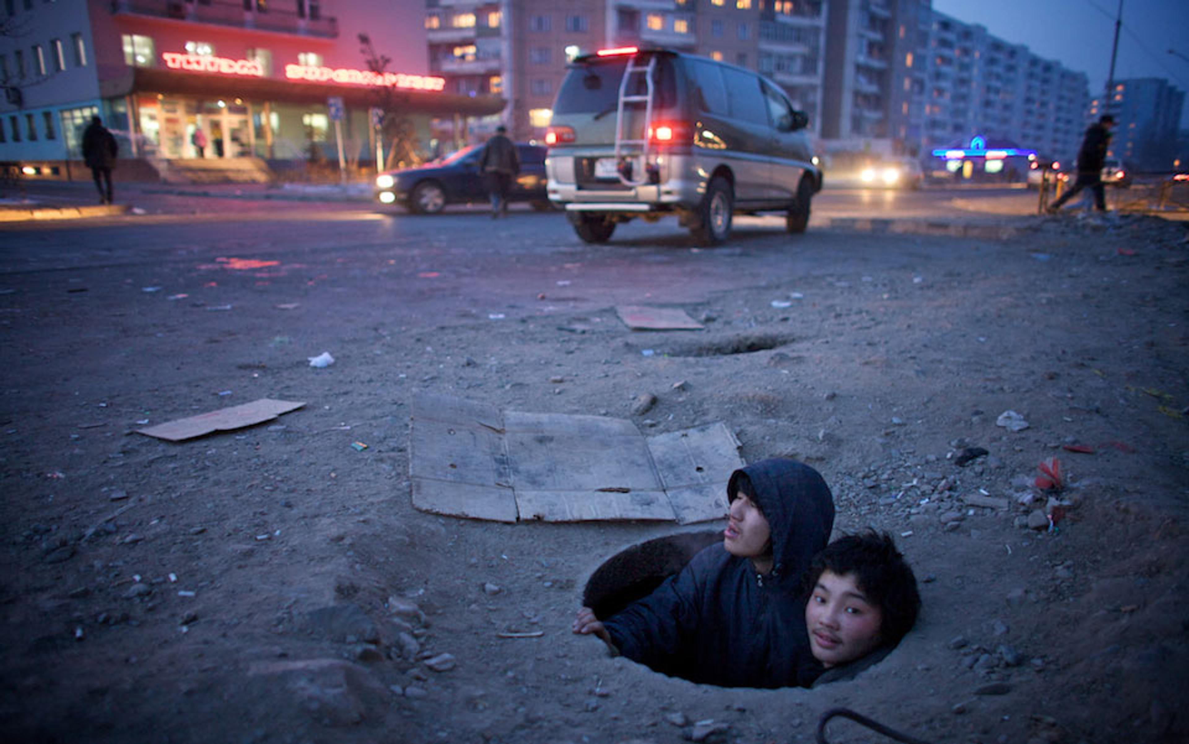Children in a city street hole at dusk with buildings, parked and moving vehicles, and a lit storefront in the background.