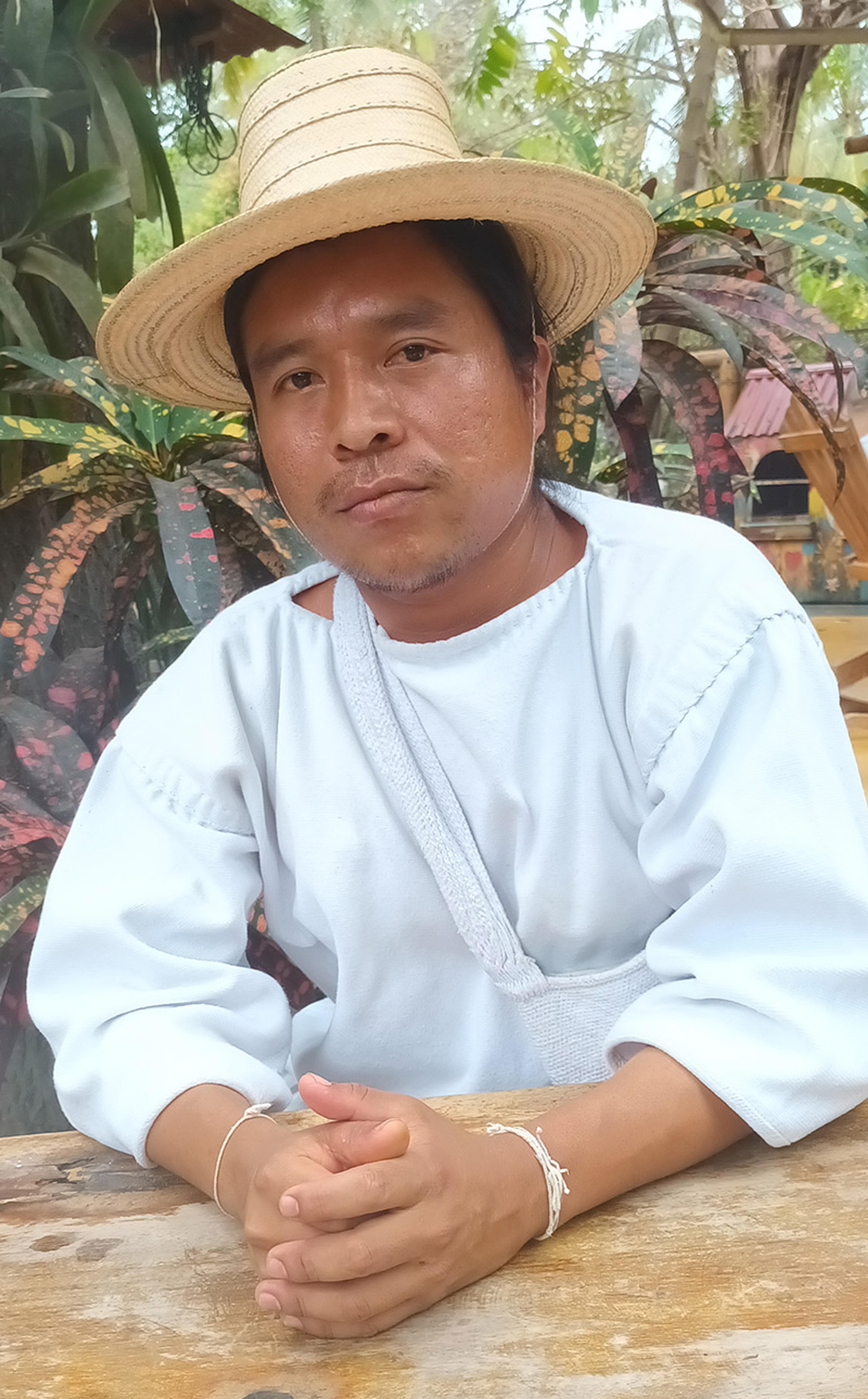 A Colombian man in a straw hat and white traditional attire, seated at a wooden table outdoors with vibrant plants in the background.
