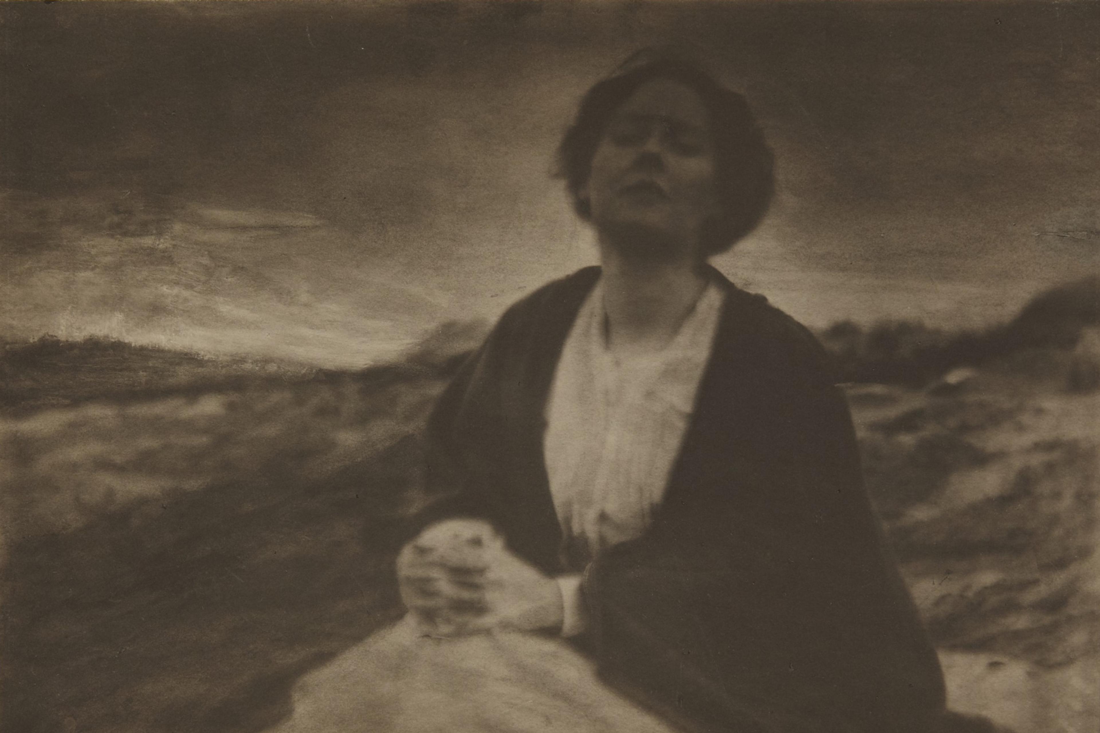 Sepia-toned photo of a woman outdoors with eyes closed and hands clasped, wearing a dark shawl against a blurred landscape.