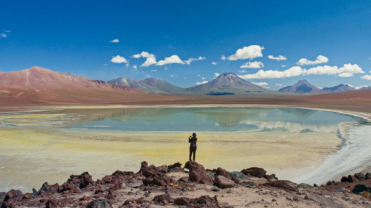 A solitary person photographing a mountain lake under a blue sky with scattered clouds in a rocky desert landscape.