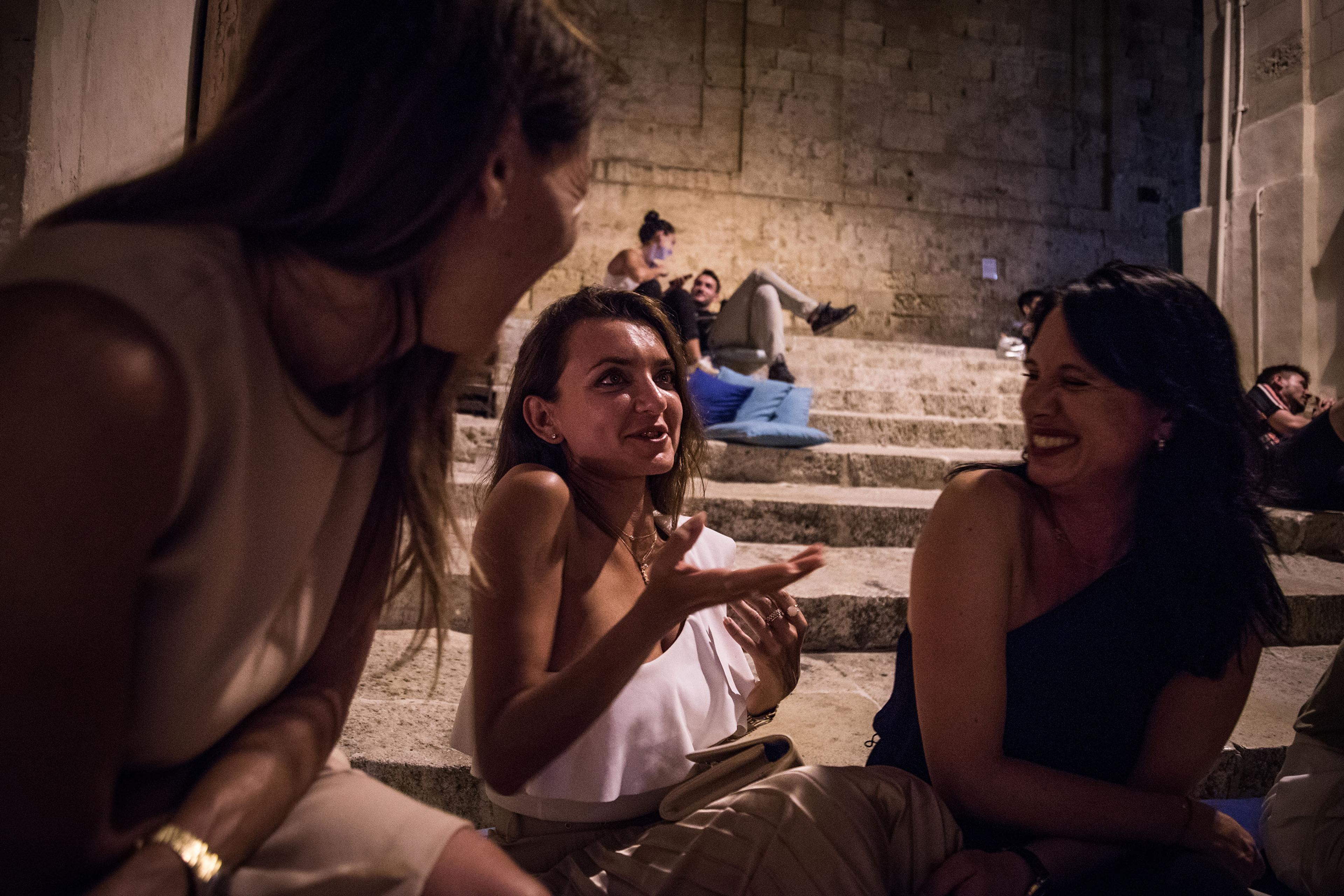 Three women chatting and laughing on stone steps at night with people relaxing in the background.