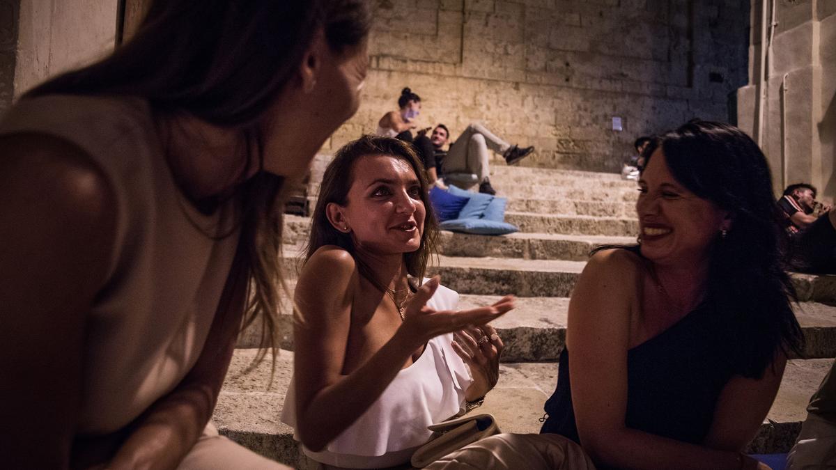 Three women chatting and laughing on stone steps at night with people relaxing in the background.