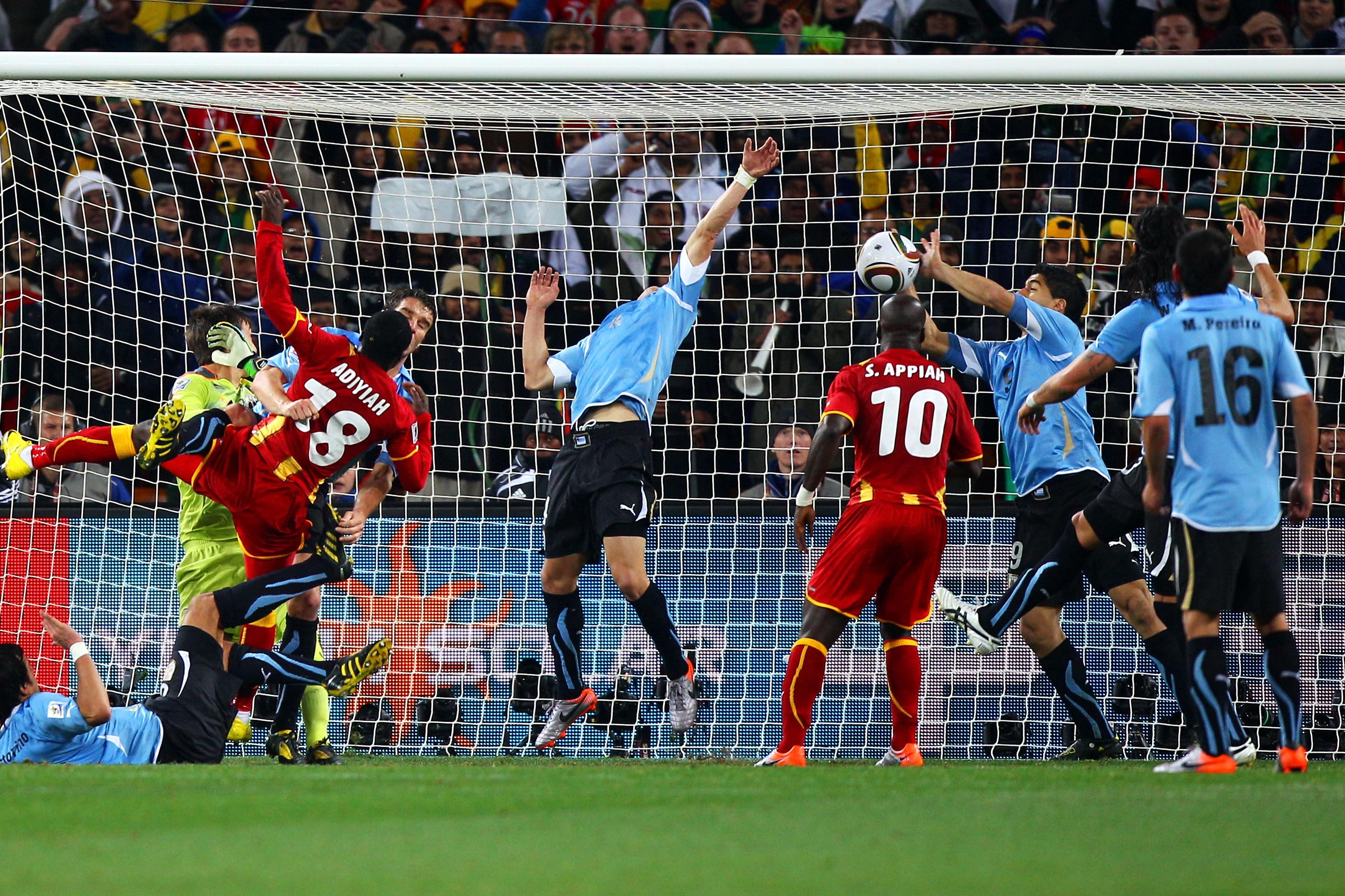 Photo of intense football match action with players crowded near the goal attempting to block or score a goal.