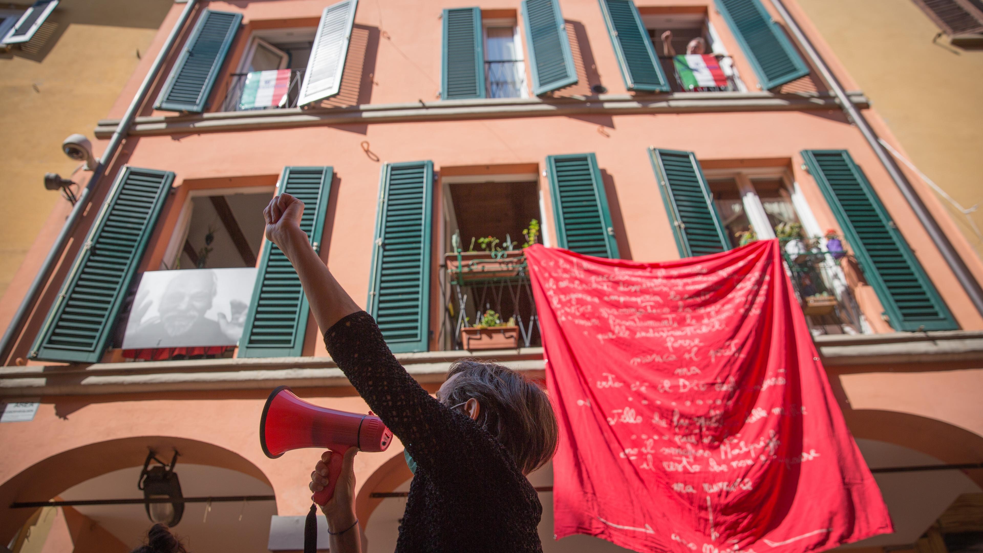 Photo of a person with a megaphone raising a fist below a building with green shutters and a red banner.