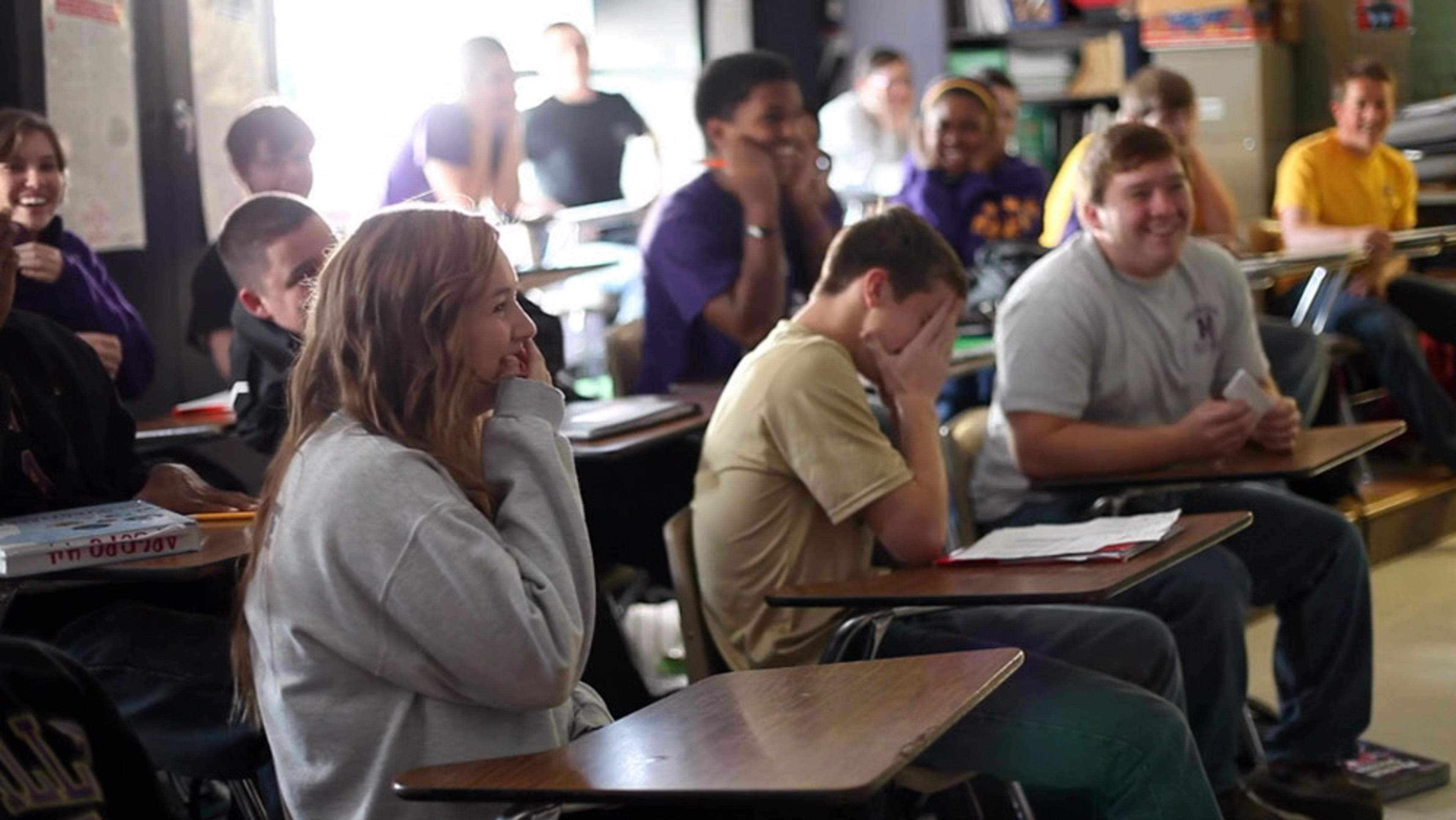 A classroom with students laughing, seated at desks, some covering their faces, casual atmosphere, sunlight entering from windows.