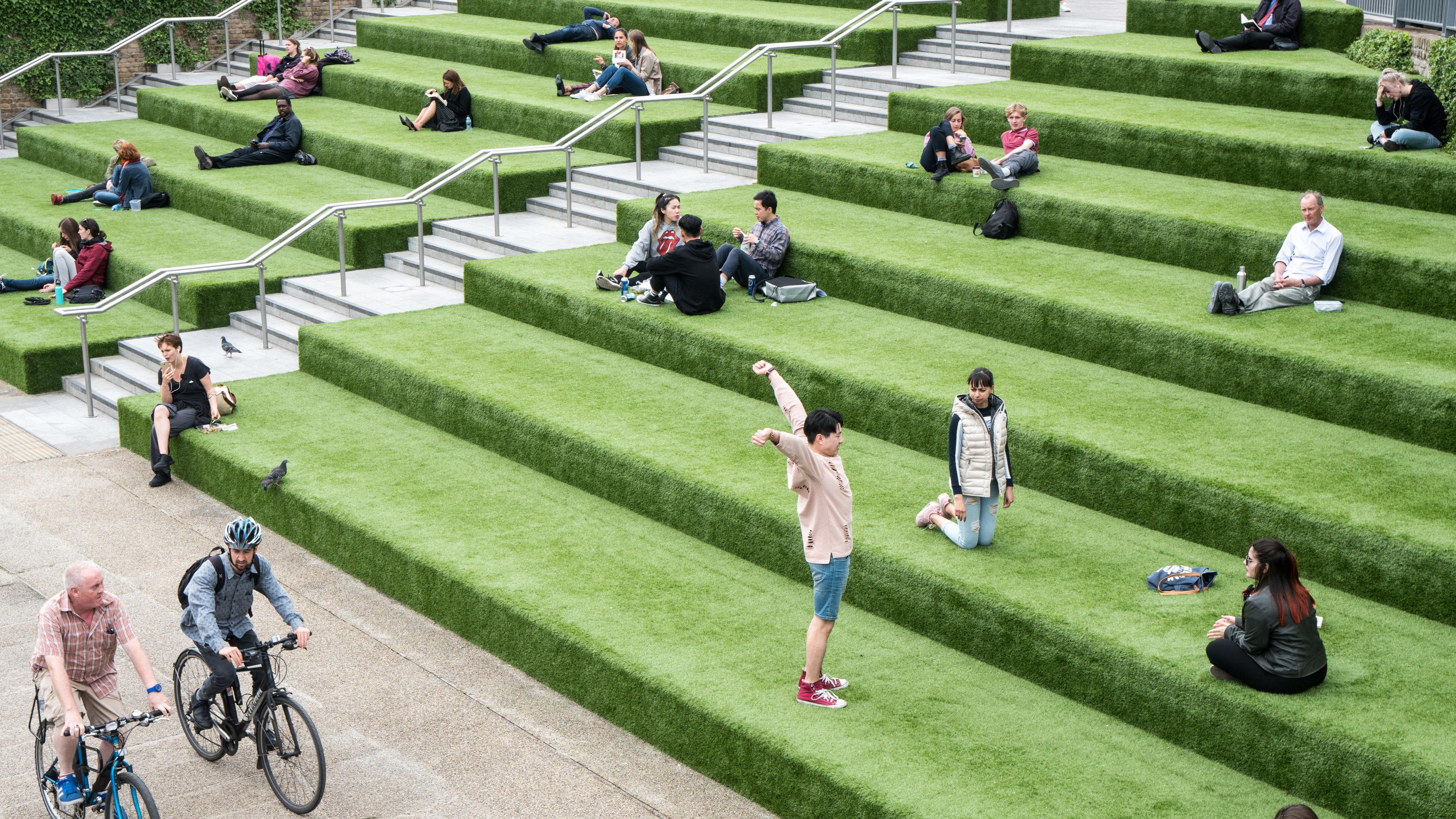 Photo of people relaxing on tiered grassy steps in a public space with railings, cyclists below, and some pigeons present.