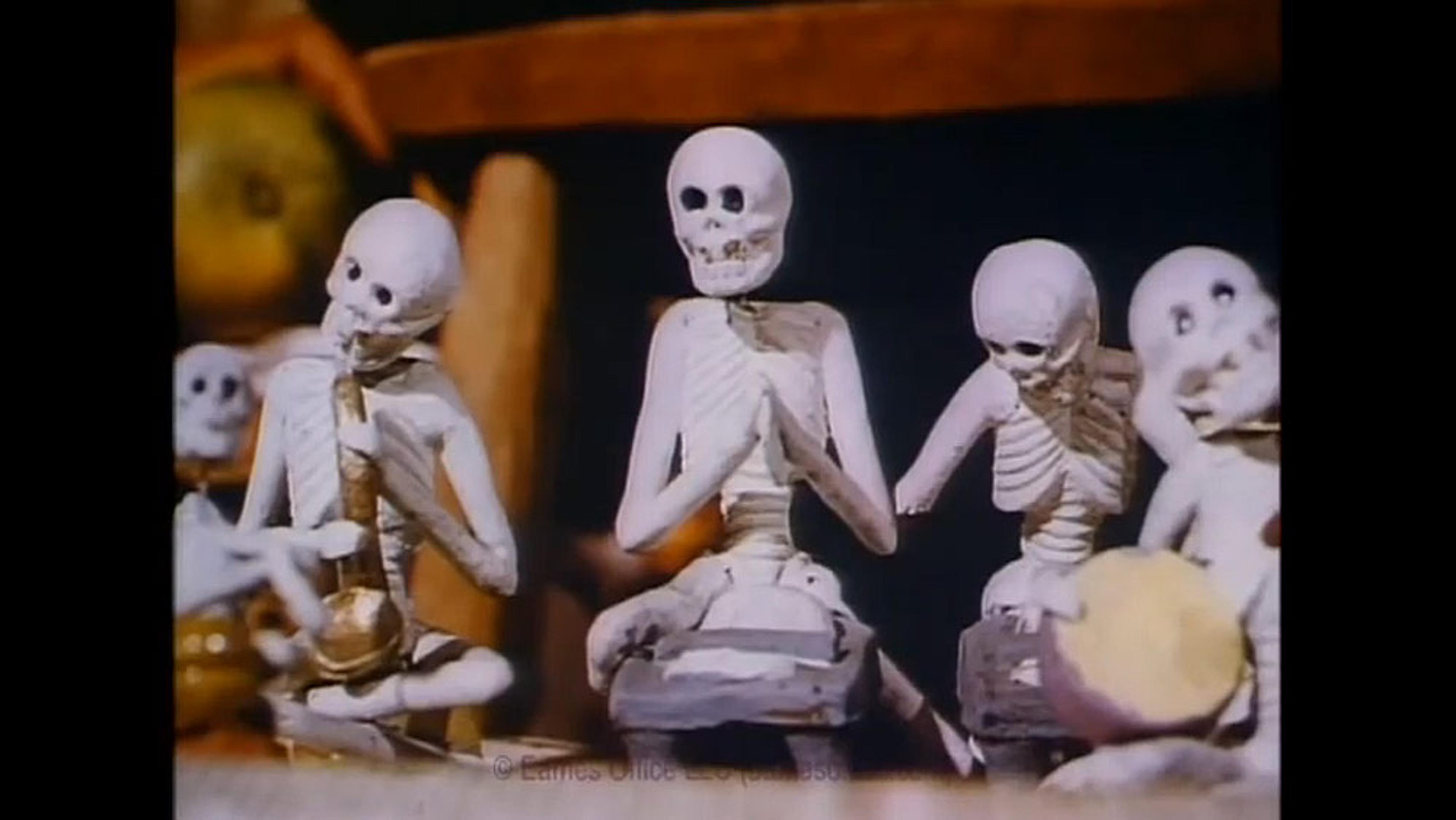 Small skeleton figures seated, playing various musical instruments, with a dark background and indistinct objects behind them.
