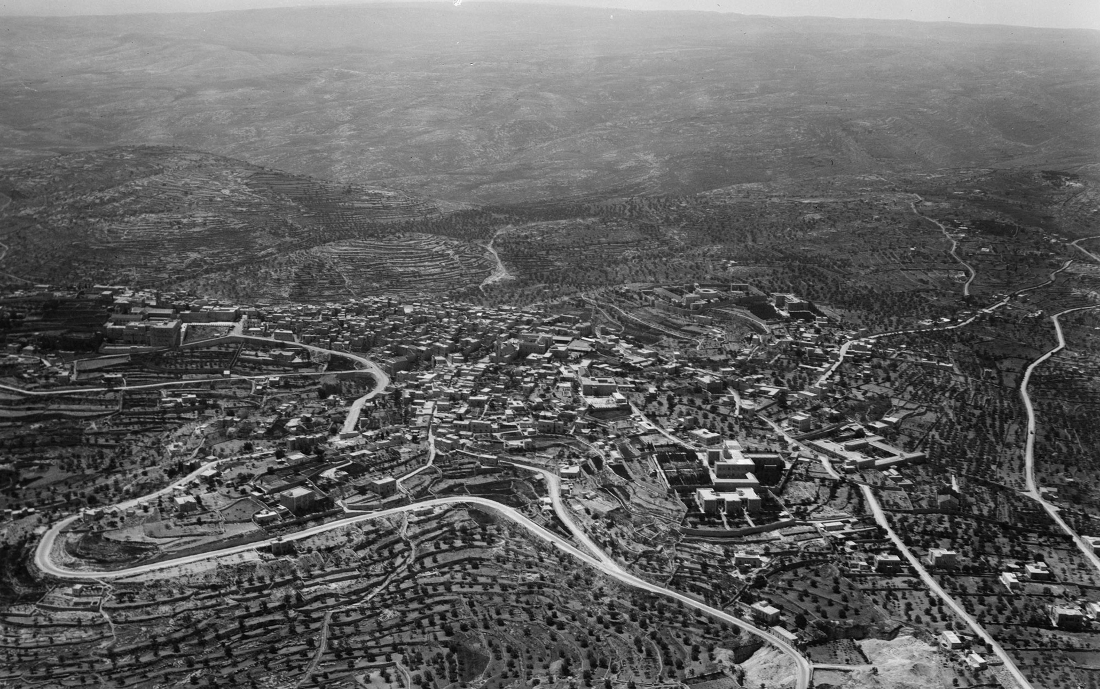 Aerial black-and-white photo of a town surrounded by rolling hills and agricultural terraces.