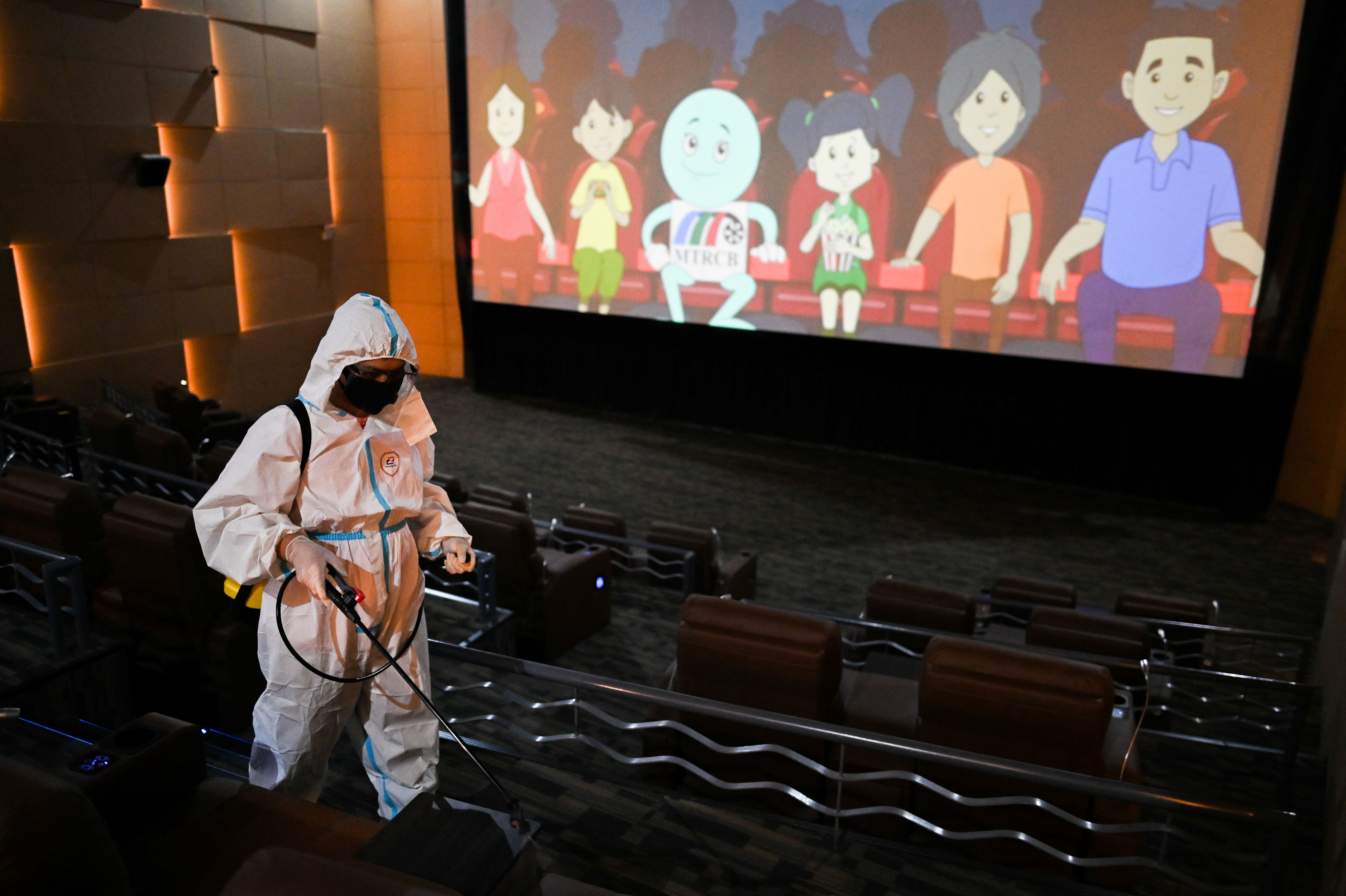 A person in protective gear disinfecting a cinema with an animated film playing on the screen in the background.