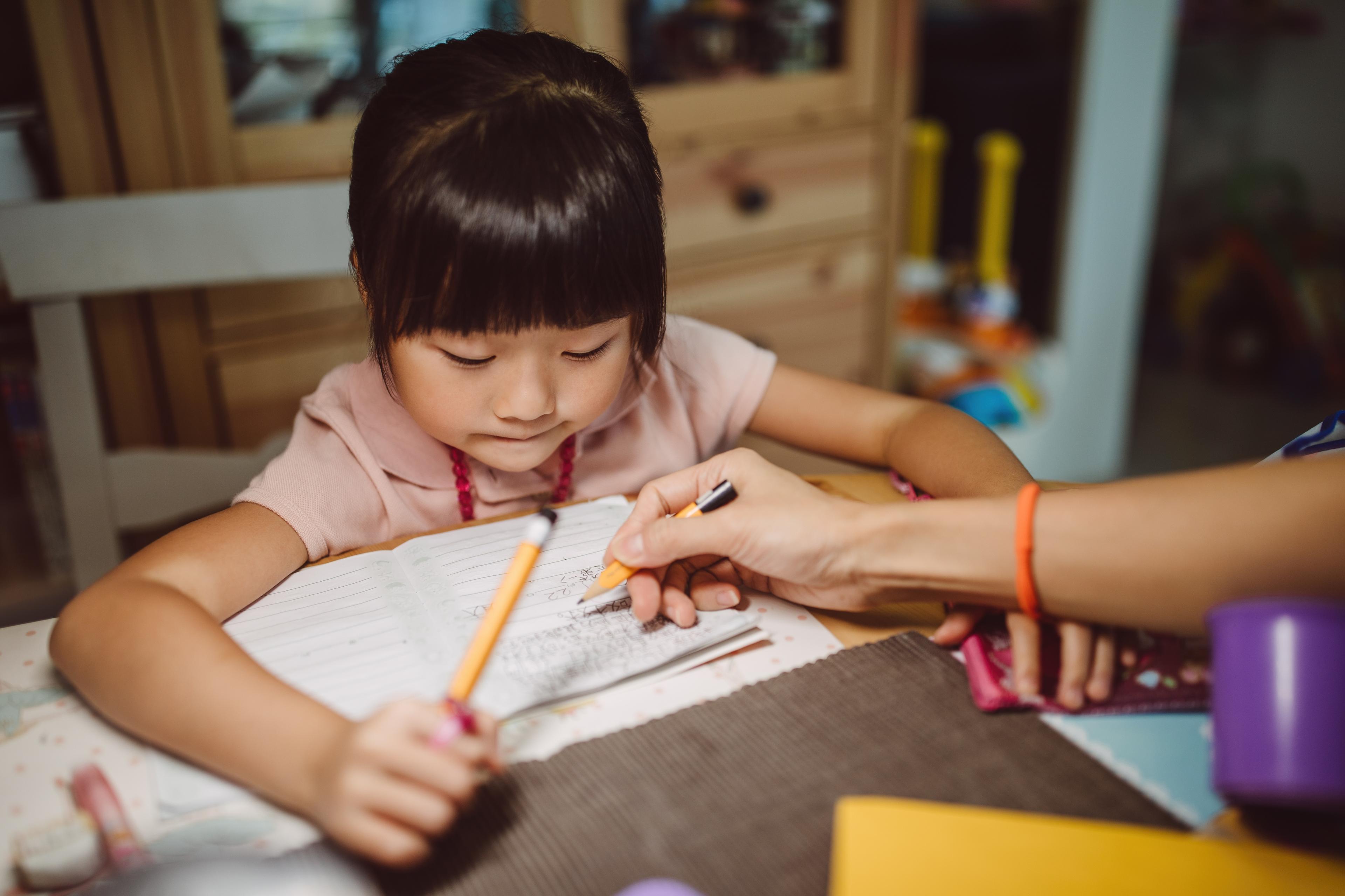 Photo of a young girl doing homework at a table with adult guidance, holding a pencil over an open notebook.