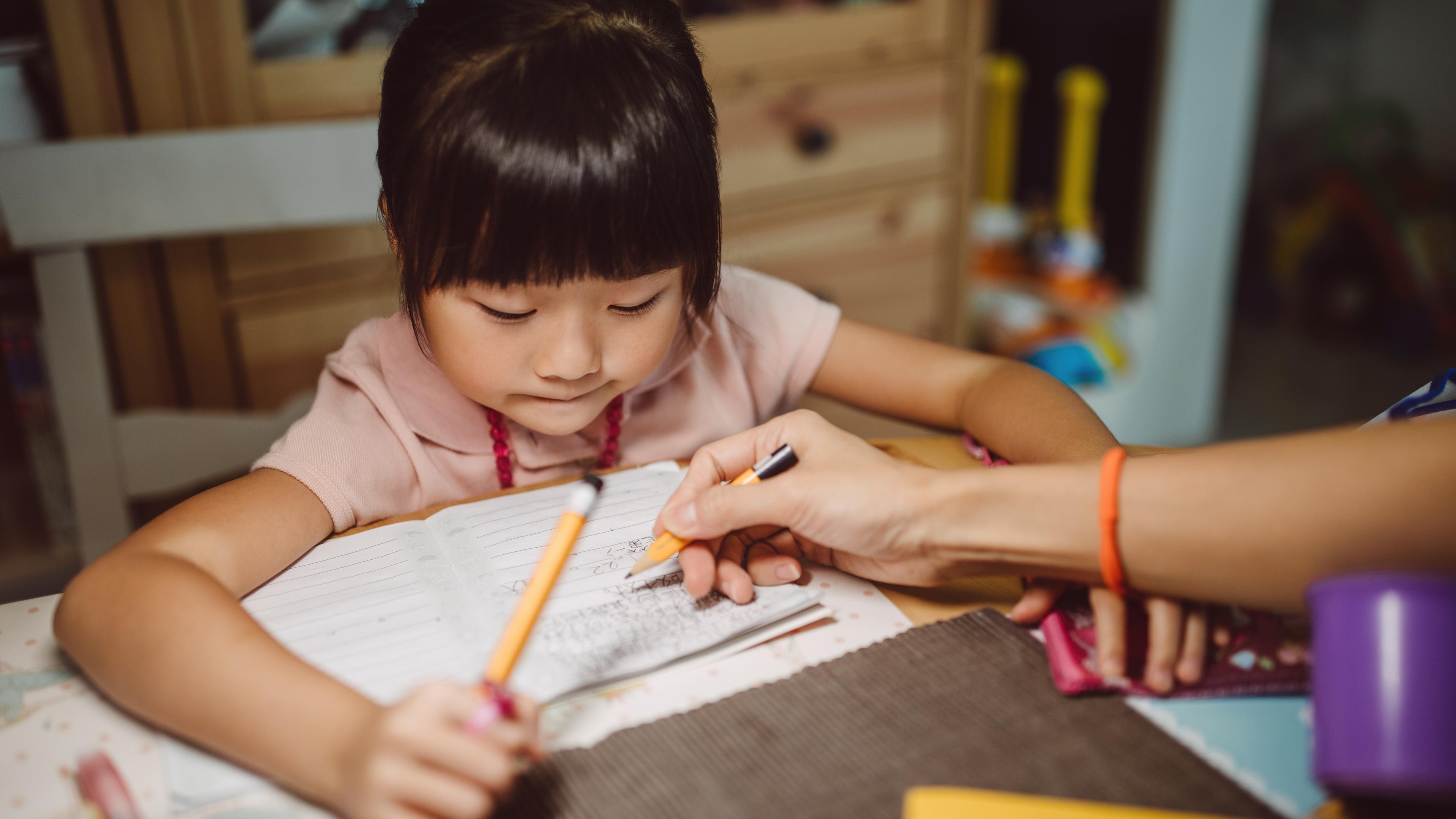 Photo of a young girl doing homework at a table with adult guidance, holding a pencil over an open notebook.