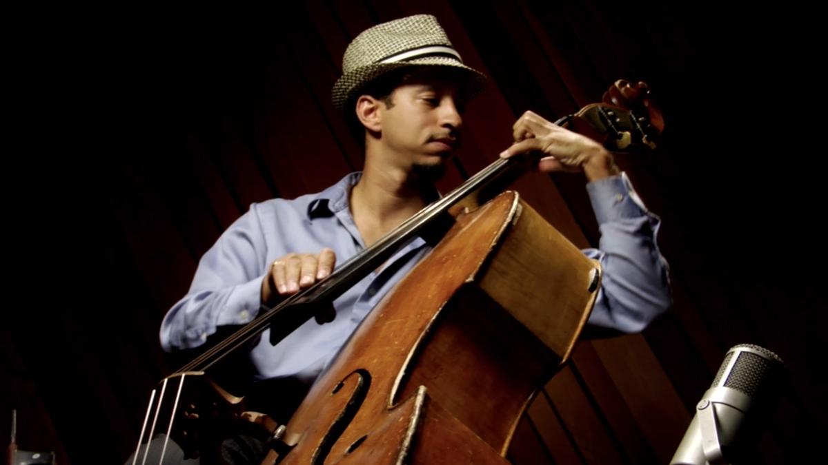 Photo of a musician in a hat playing a double bass with a microphone in front, set against a dark background.