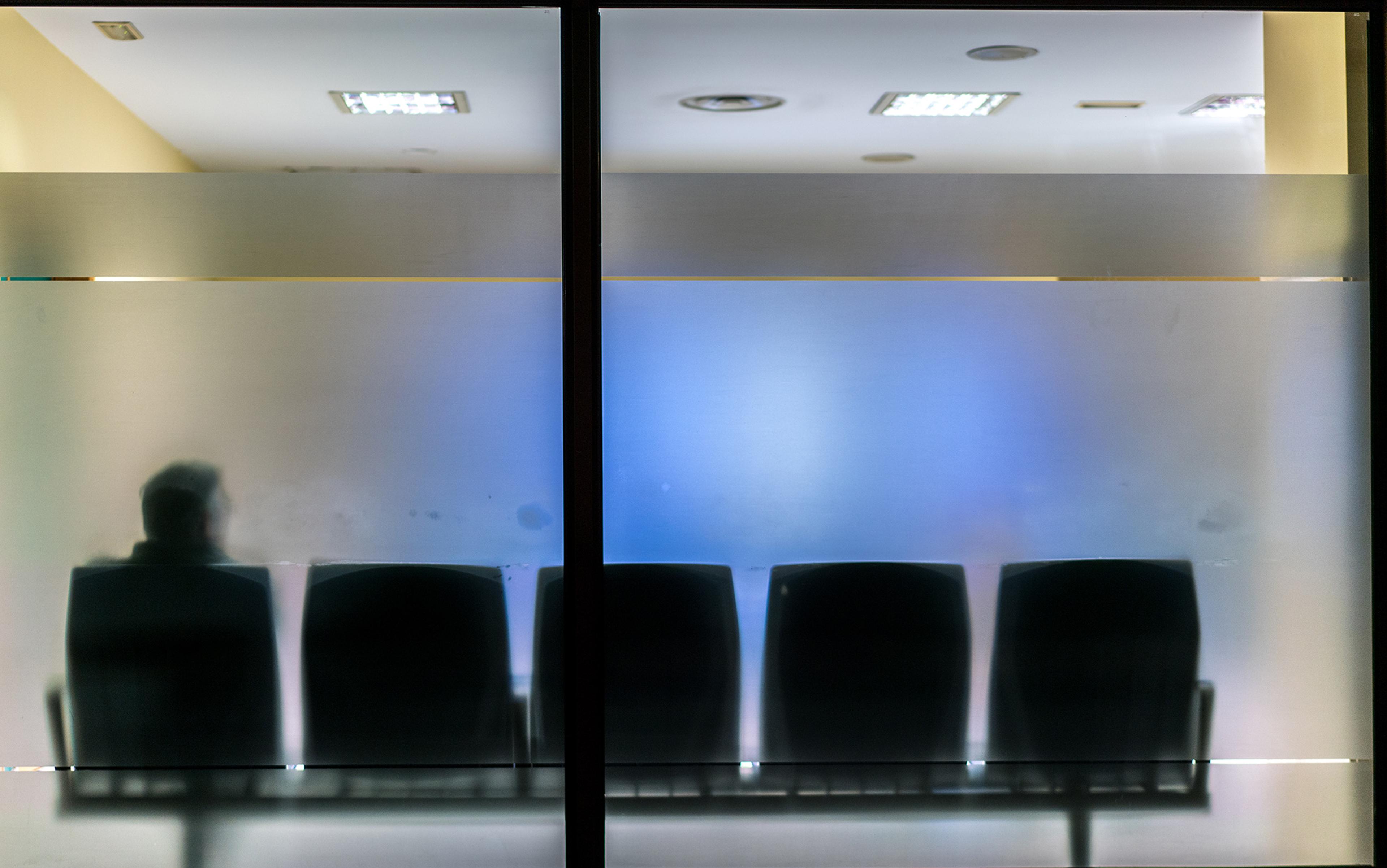 A silhouetted figure sitting on a chair behind frosted glass partitions in a waiting room with overhead lights.