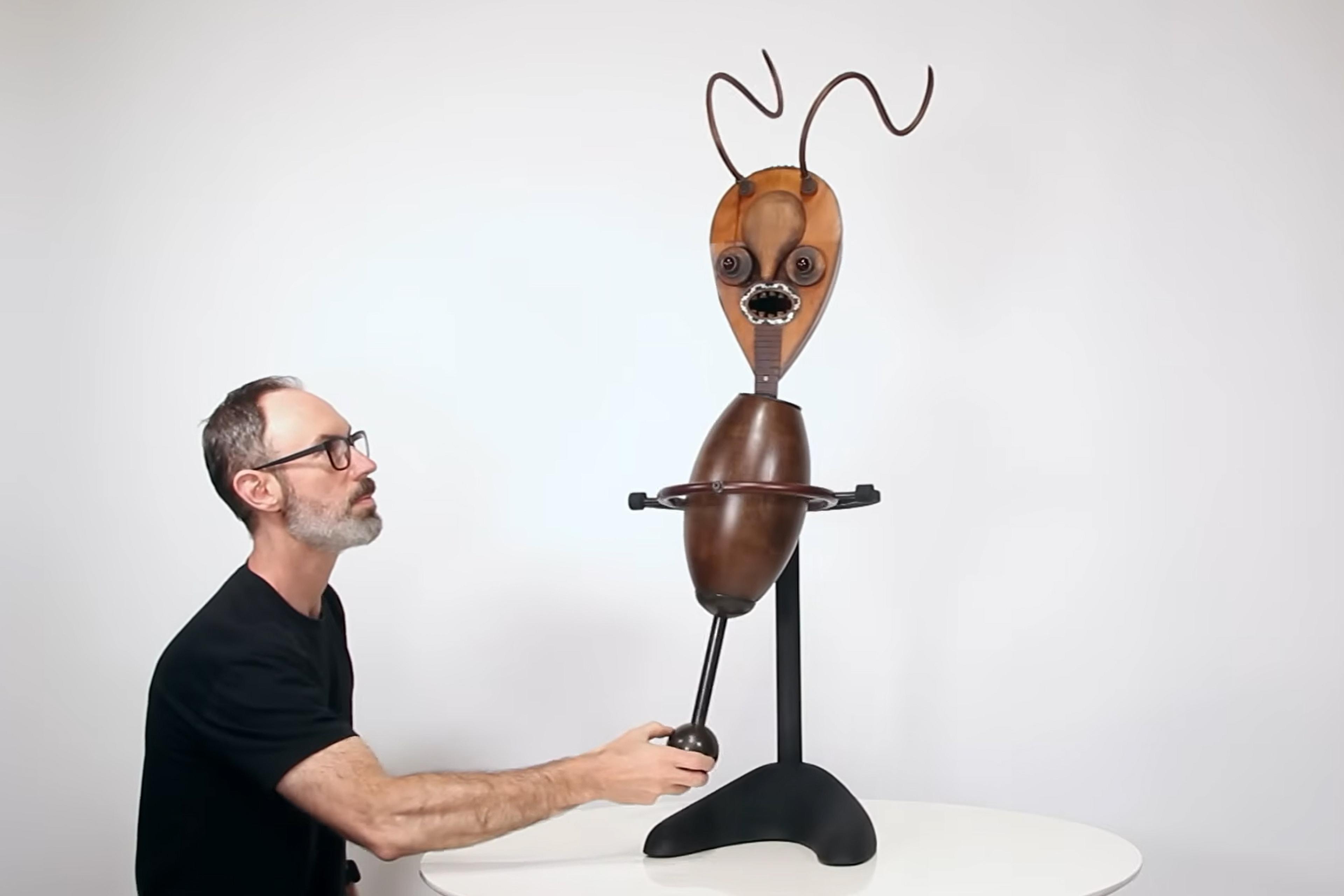 A man interacts with a quirky wooden sculpture with antler-like wires on a table against a plain background; photo.