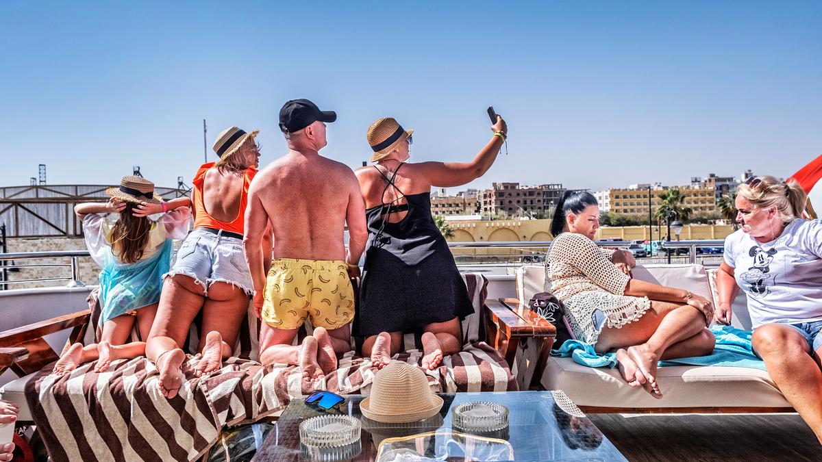 People on a boat deck taking a selfie against a city skyline with clear blue sky, towels and sun hats visible.